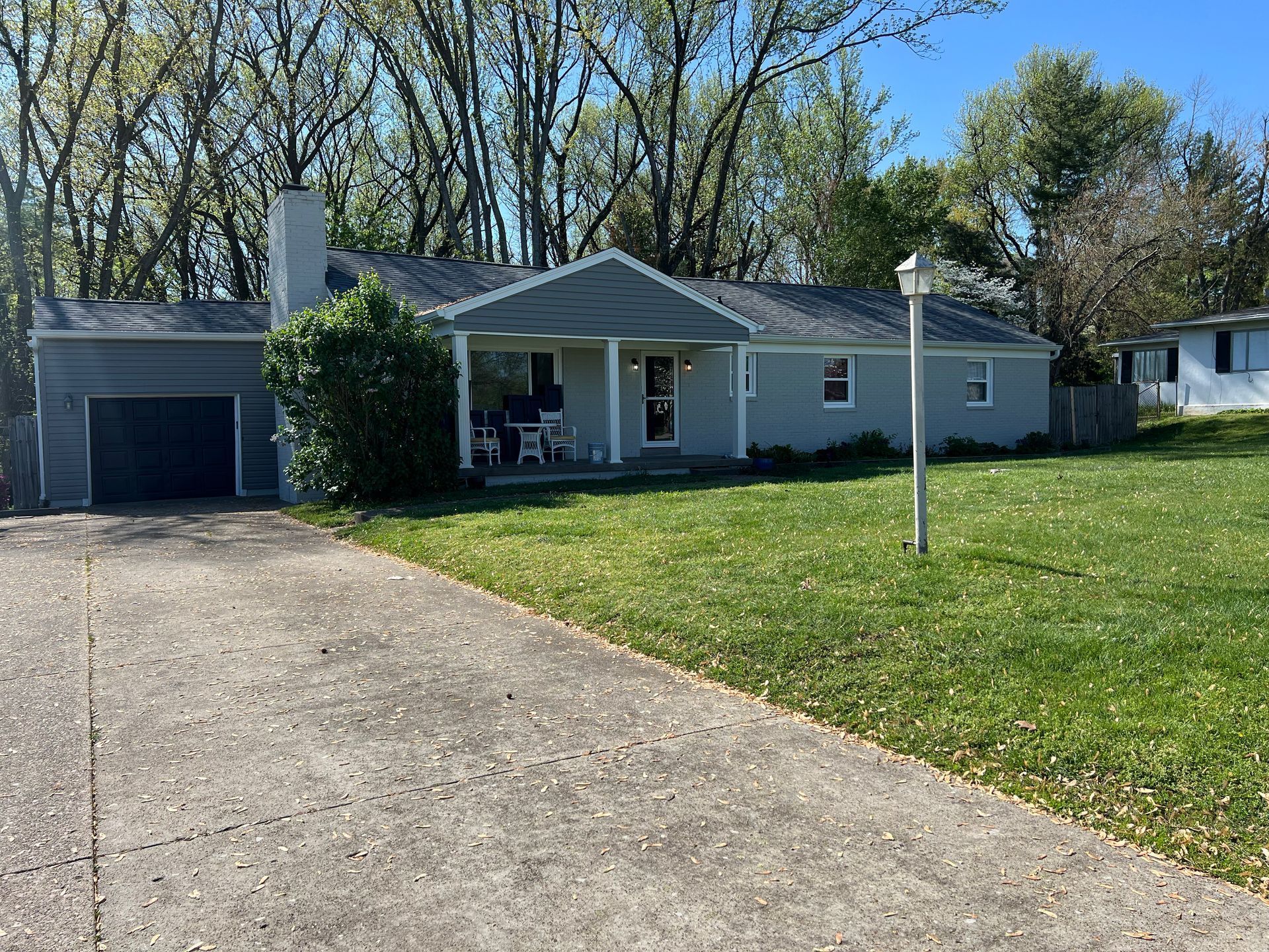 Gray ranch-style house with a driveway, garage, and front porch. Green lawn and trees in the background on a sunny day.