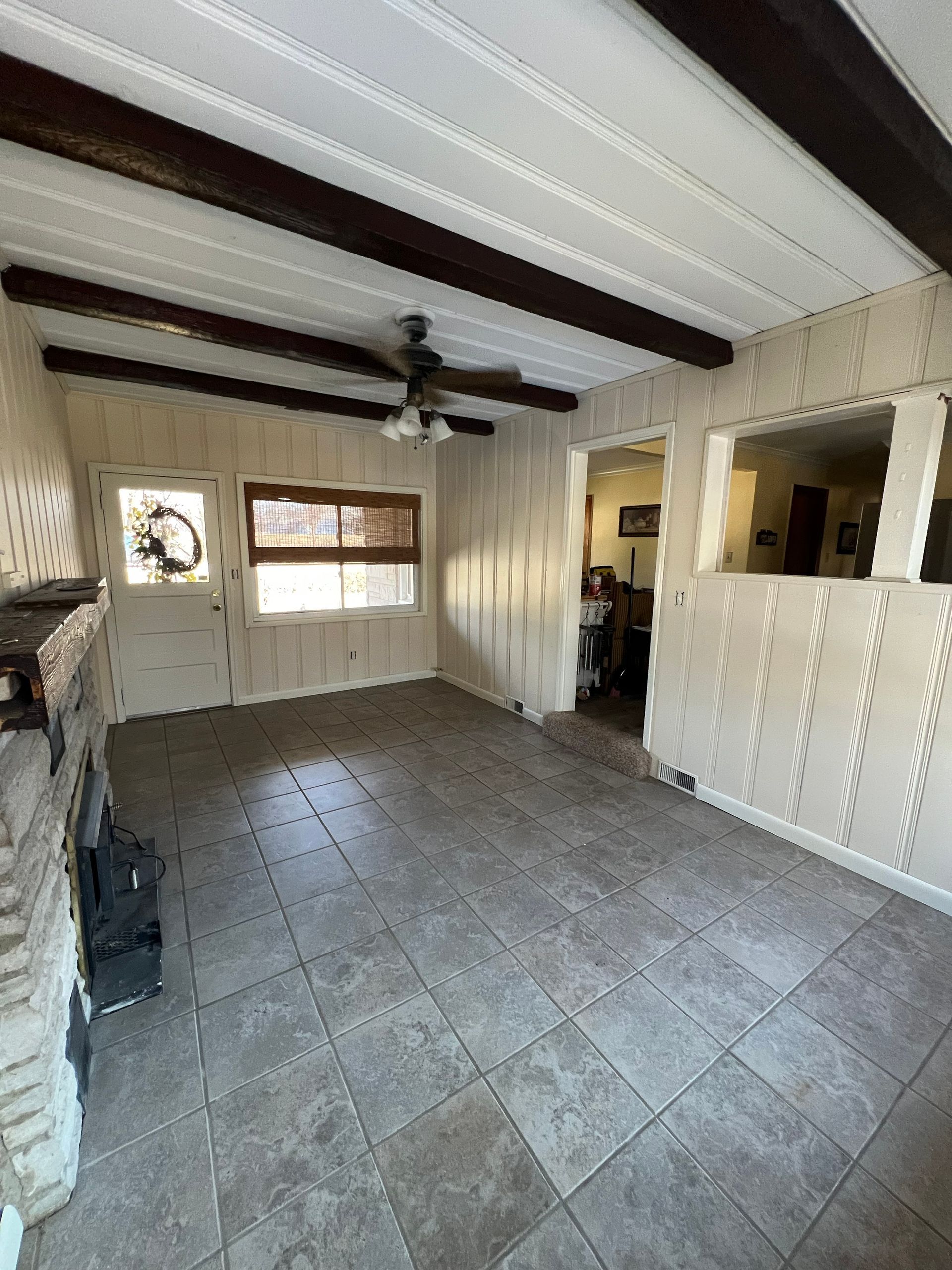 Empty living room with tile floor, paneled walls, dark beams, and a brick fireplace.