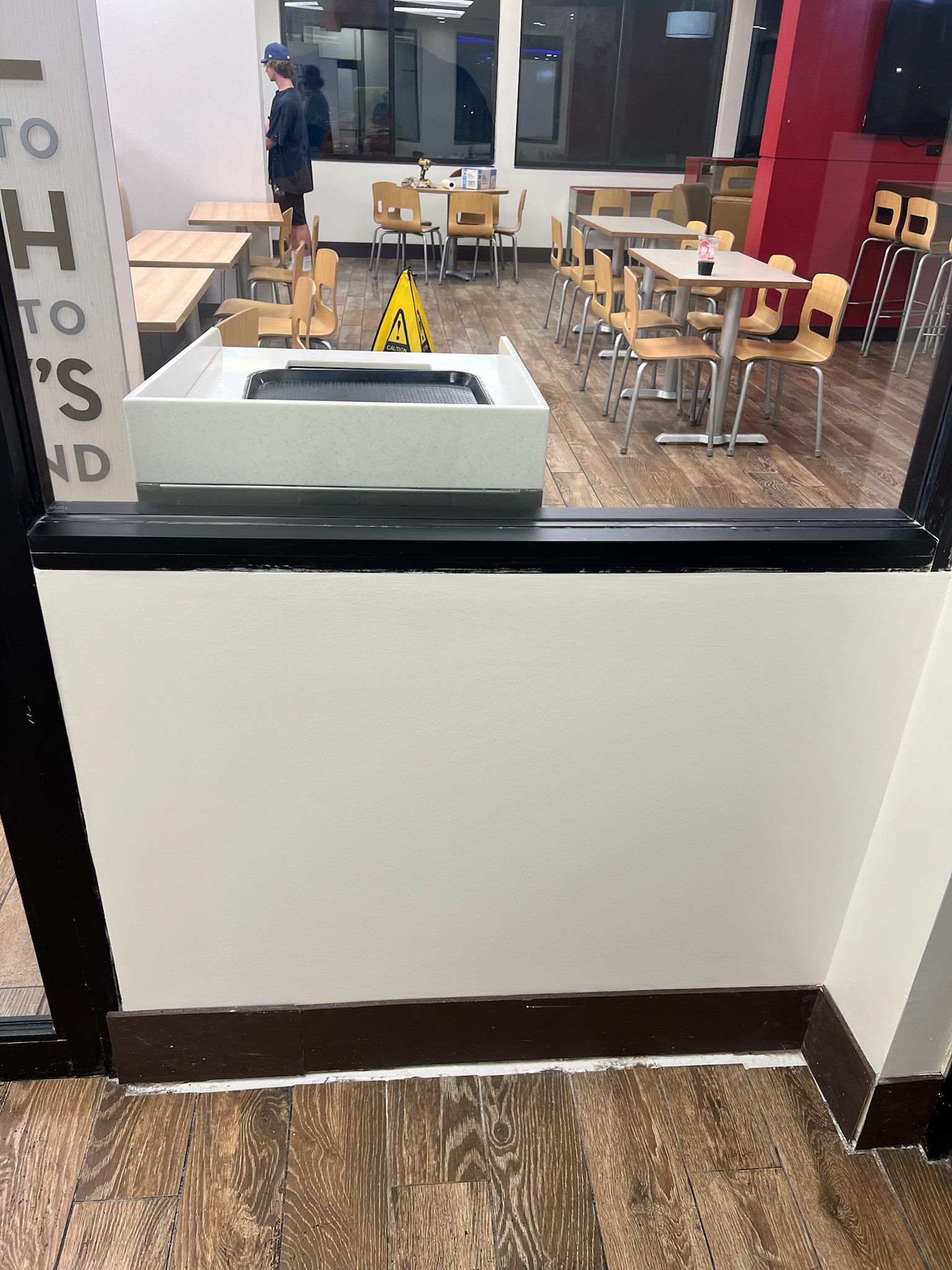 A view through a glass window, showing an empty restaurant with tables, chairs, and a trash receptacle.