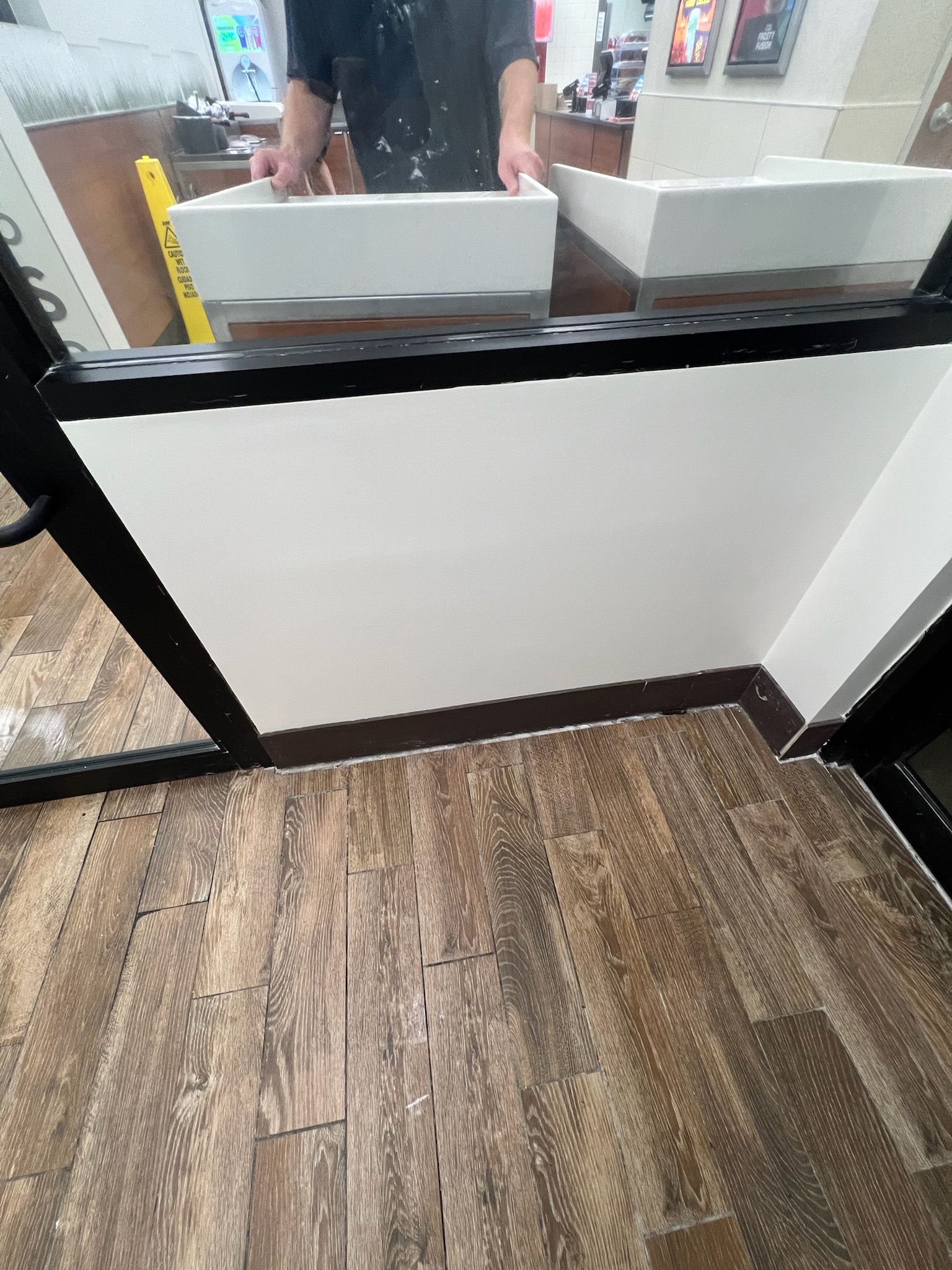 Brown wood-look floor, white wall, black door frame. Person holding white sinks is reflected in glass.