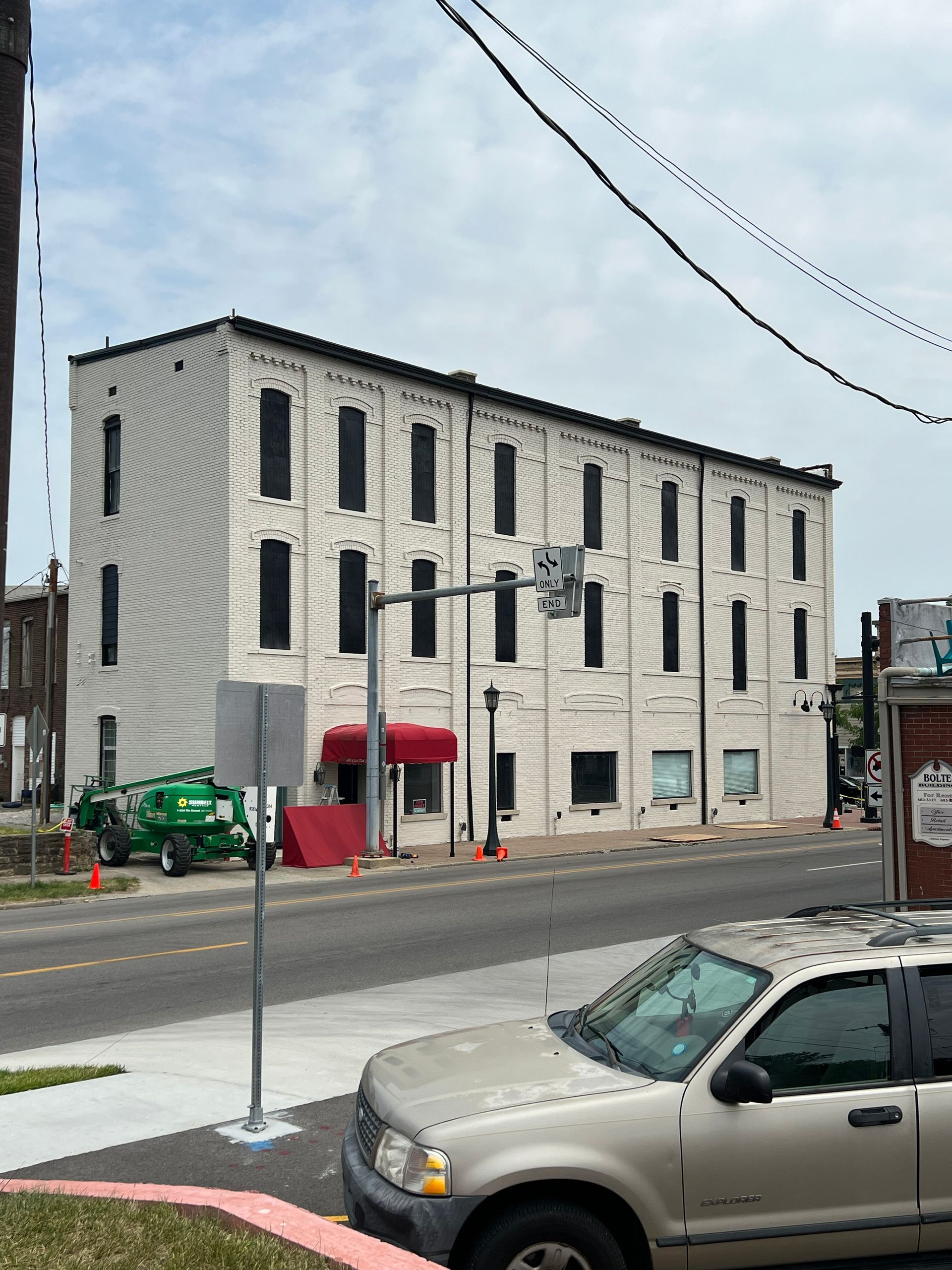 White brick building with black windows and a red awning on a city street. A silver car and a green vehicle are in the foreground.