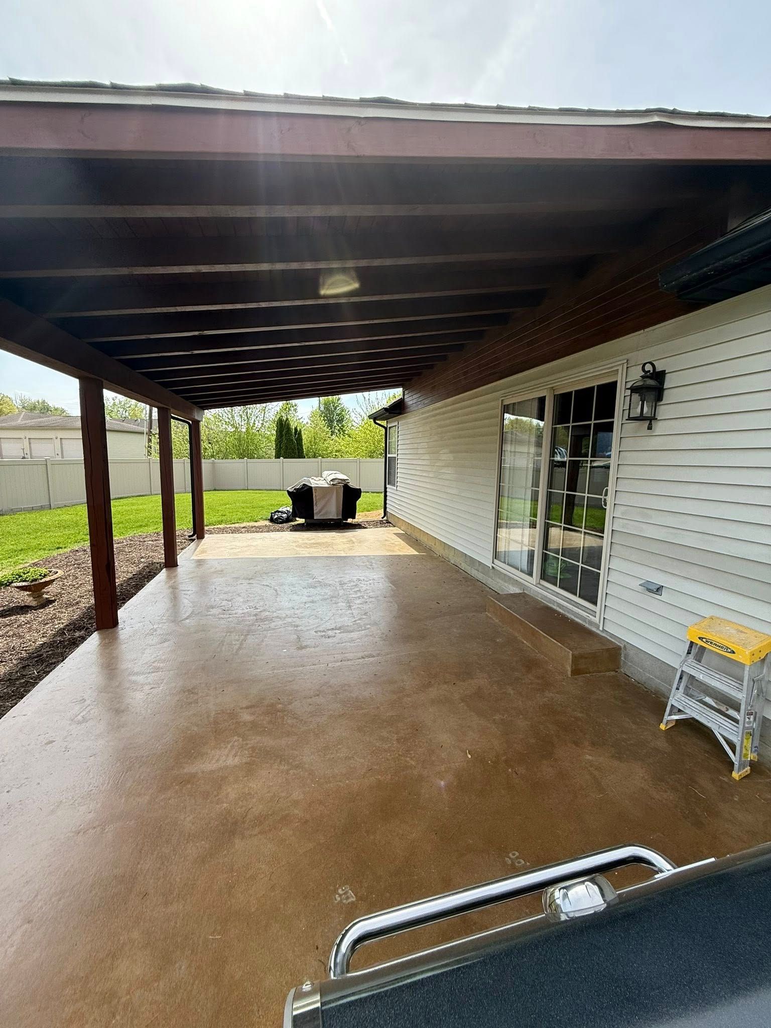 Covered patio with brown beams, concrete floor, white siding, and a grill in the background.