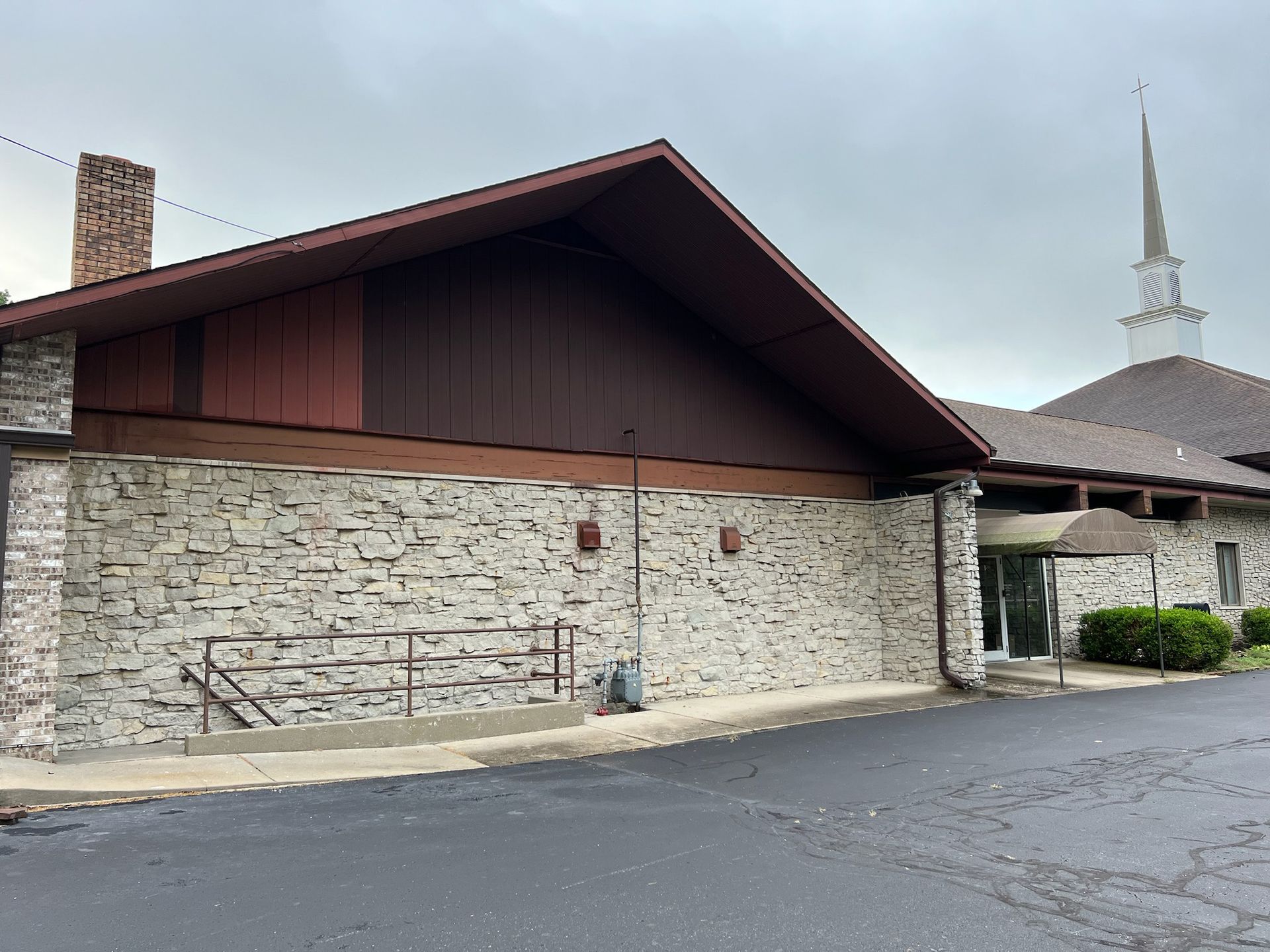 A church building with stone walls and a brown roof. A steeple is visible in the background.