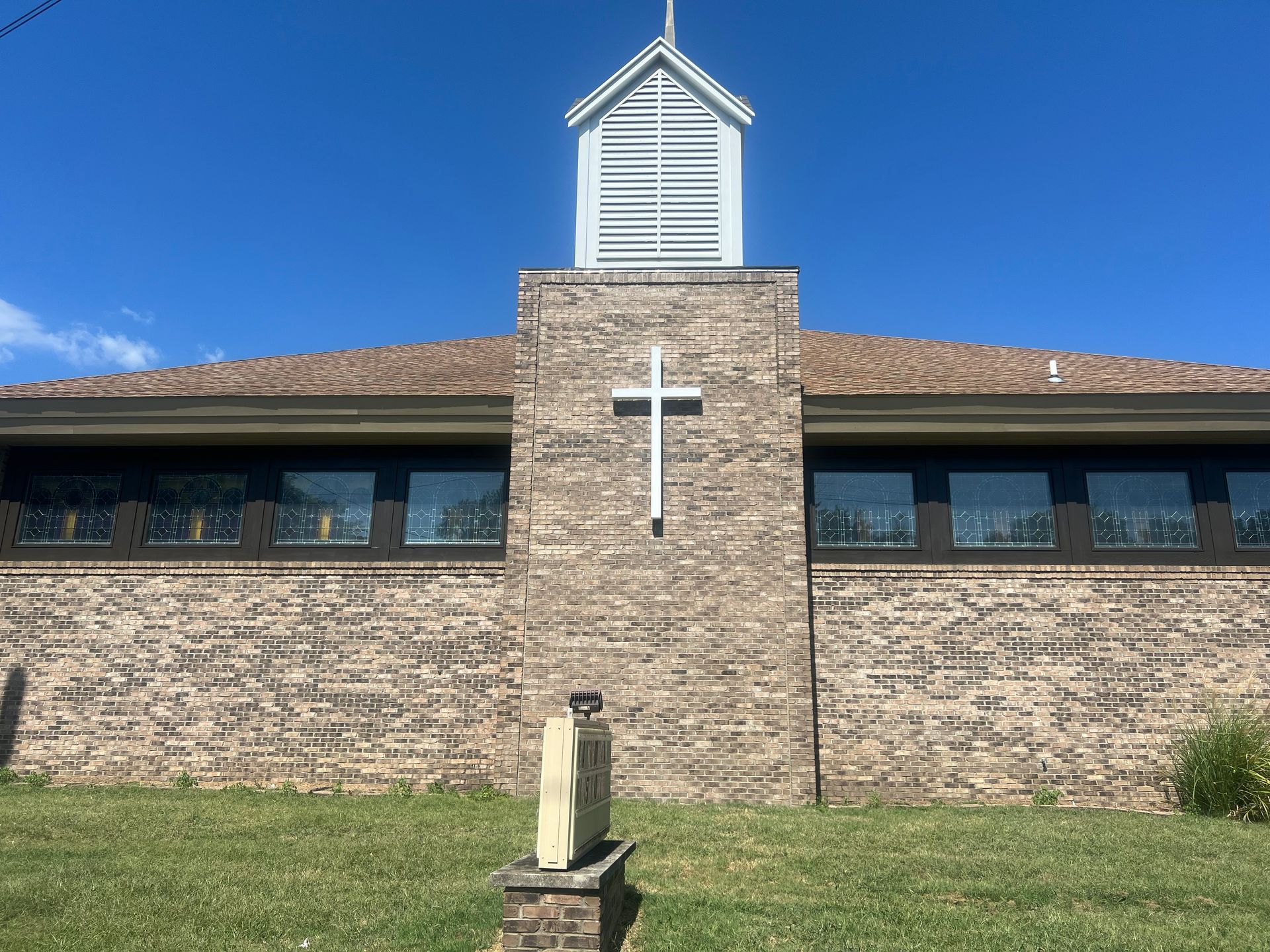 Church with brick facade, white cross, steeple, and stained-glass windows. Clear blue sky.