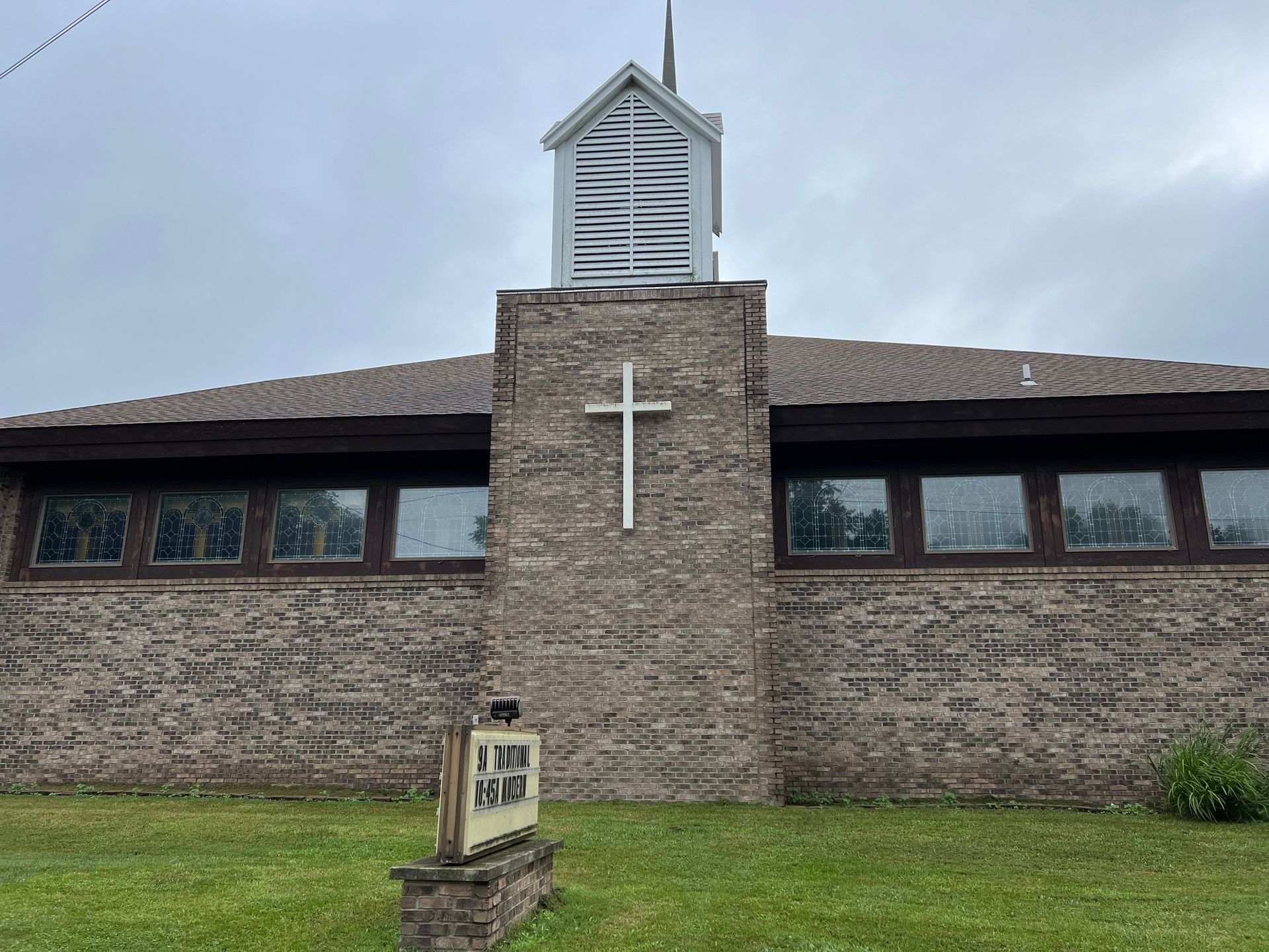 A church building with a white cross, a bell tower, and a sign in front, under a cloudy sky.