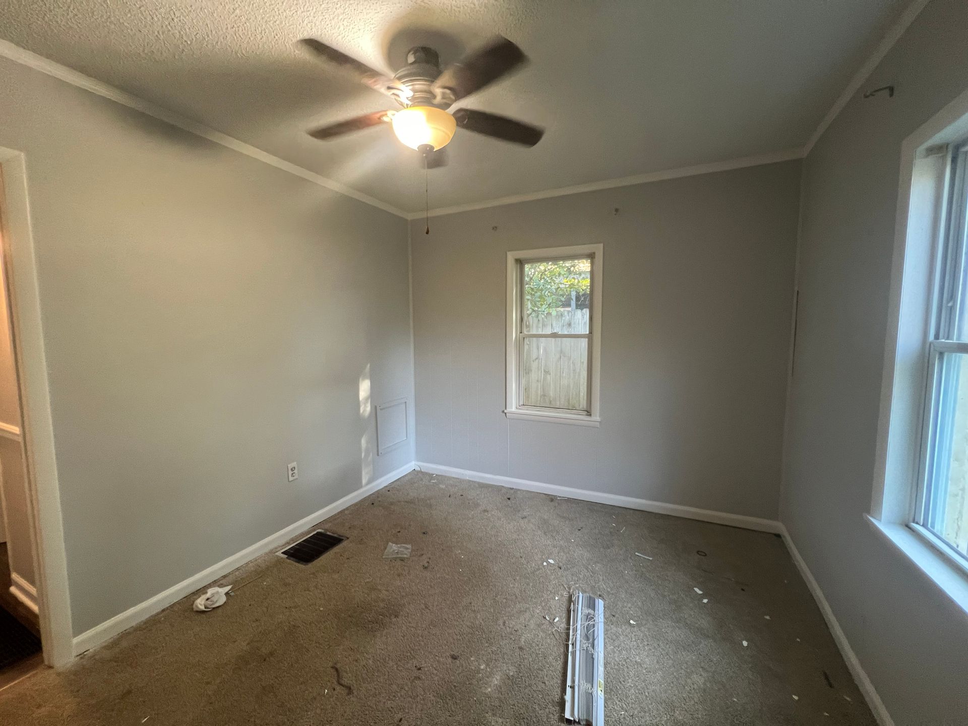 Empty room with stained carpet, grey walls, ceiling fan, and two windows.