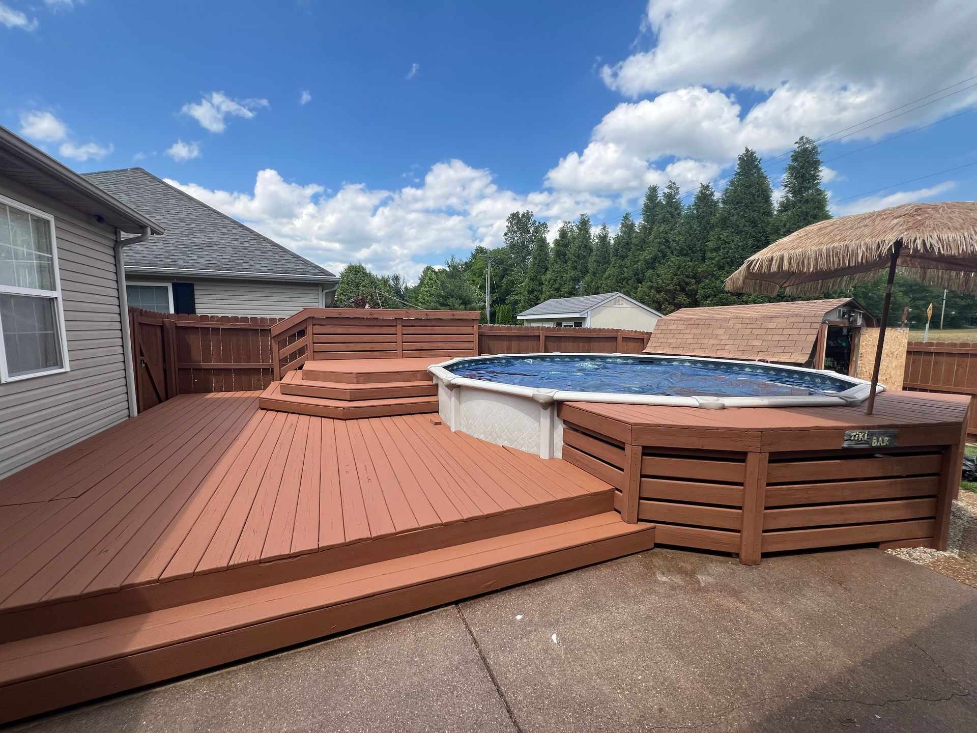 Brown deck with above-ground pool. Stairs lead up to a deck area. Sunny day.