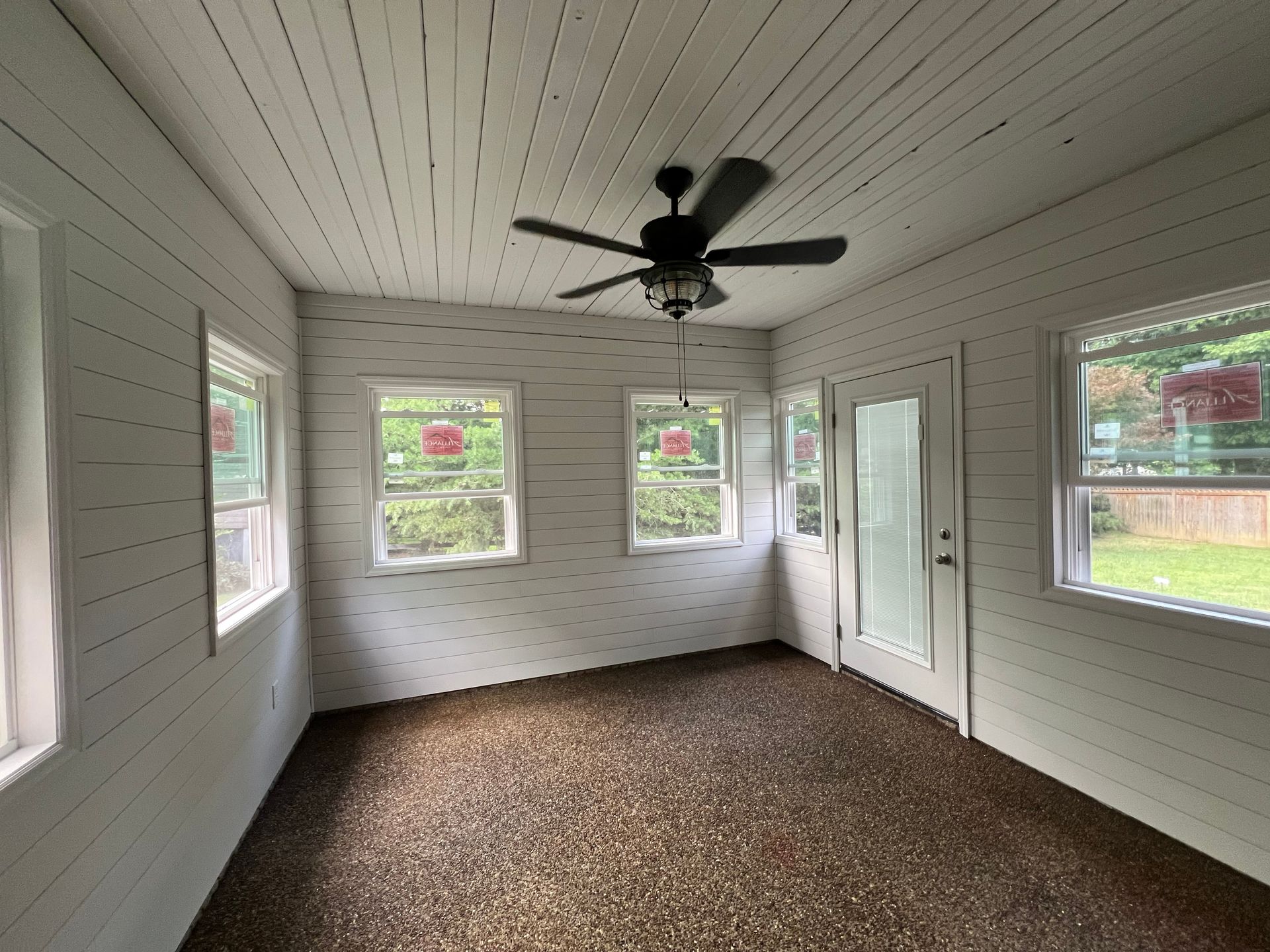 Sunroom with white plank walls and ceiling, windows, a door, and a ceiling fan; brown pebble floor.