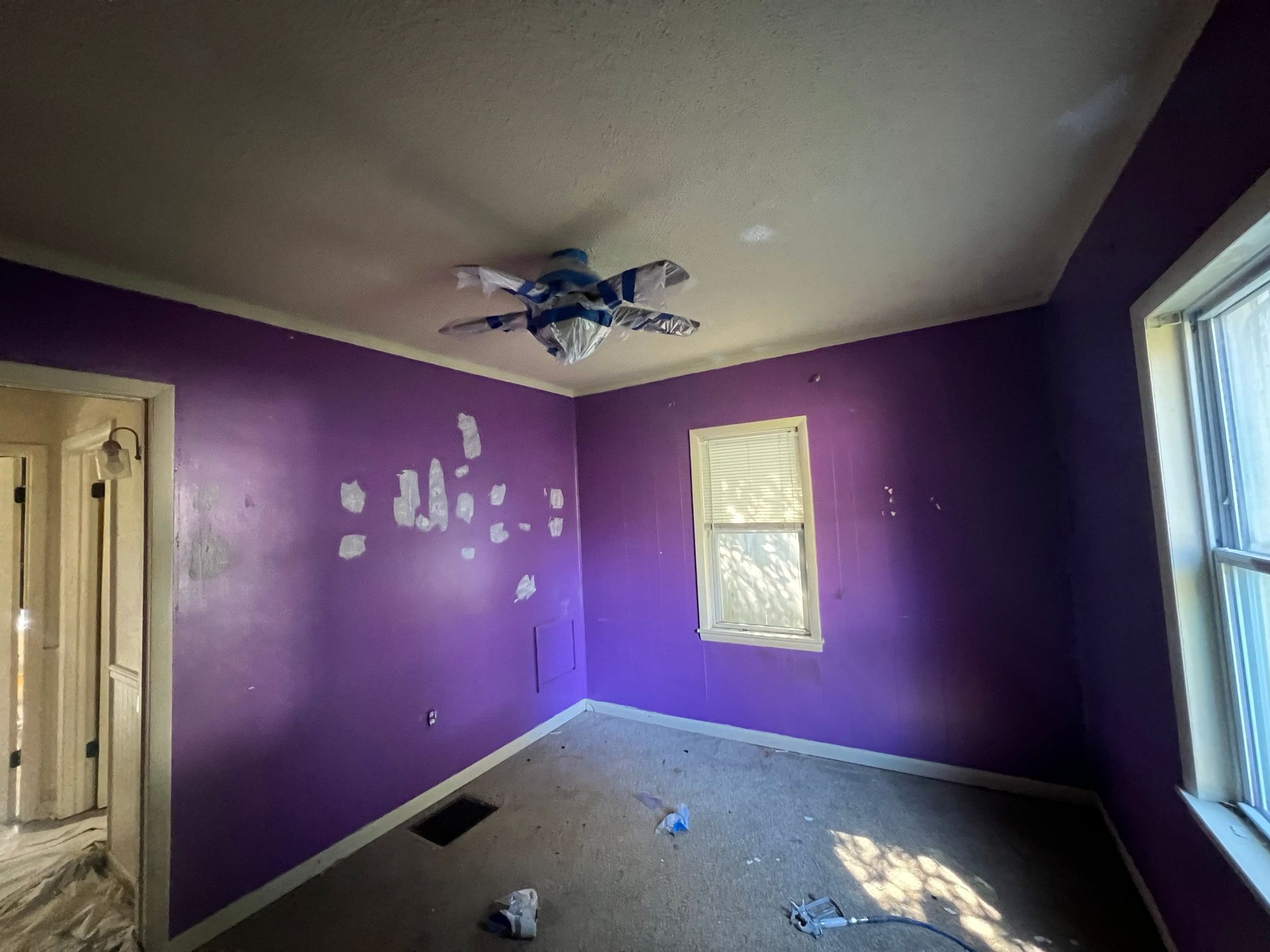 Purple bedroom with patched walls, ceiling fan, small window, and a doorway.