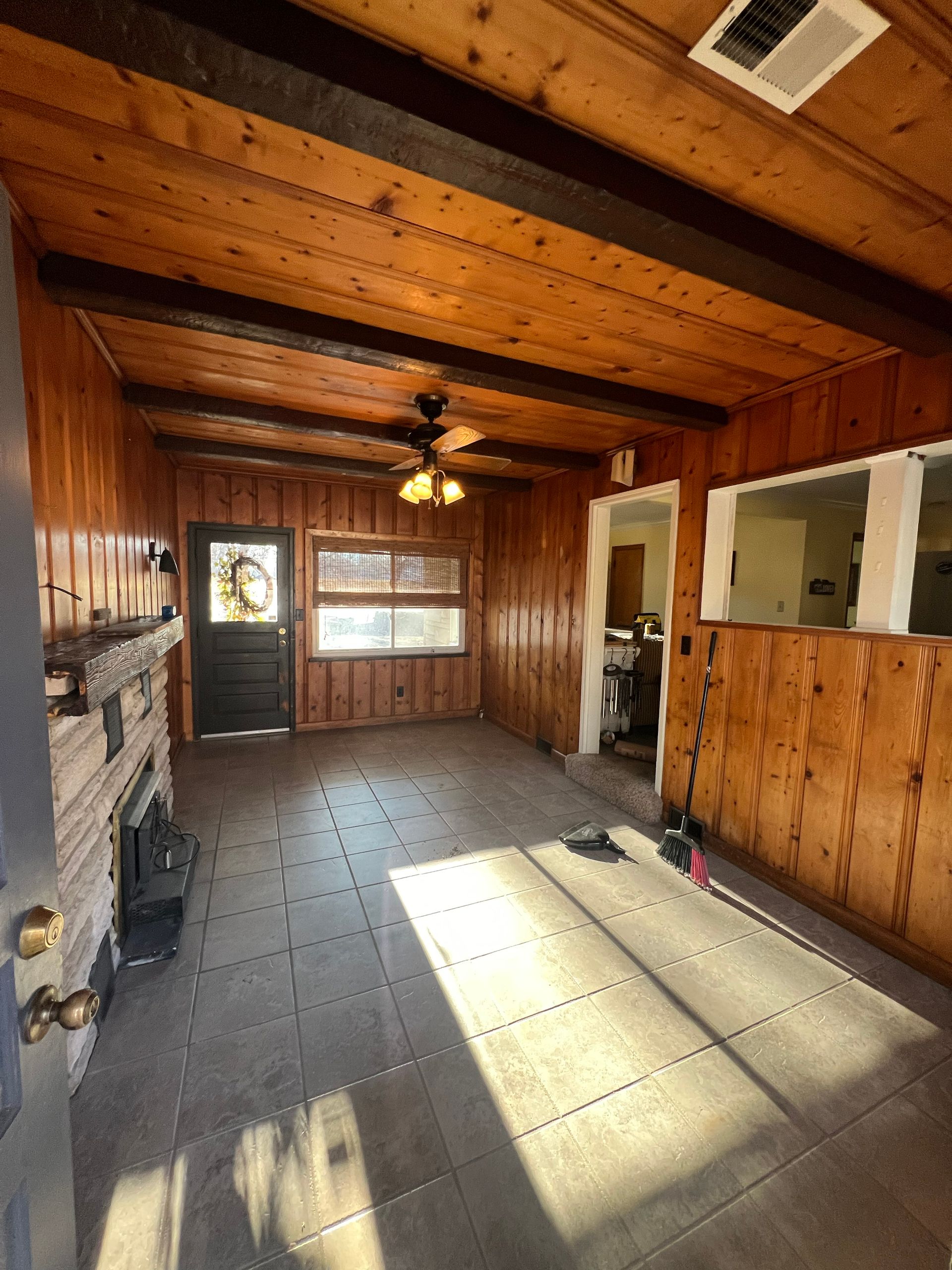 Wood-paneled room with a fireplace, tile floor, and a door. Sunlight streams in from a window.