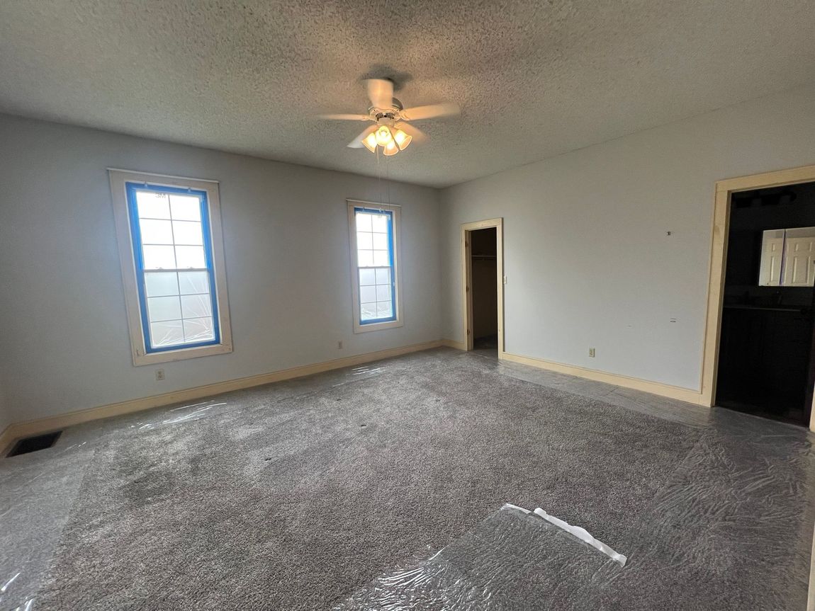 Empty room with gray walls, two windows with blue tape, dark carpet, and a ceiling fan.