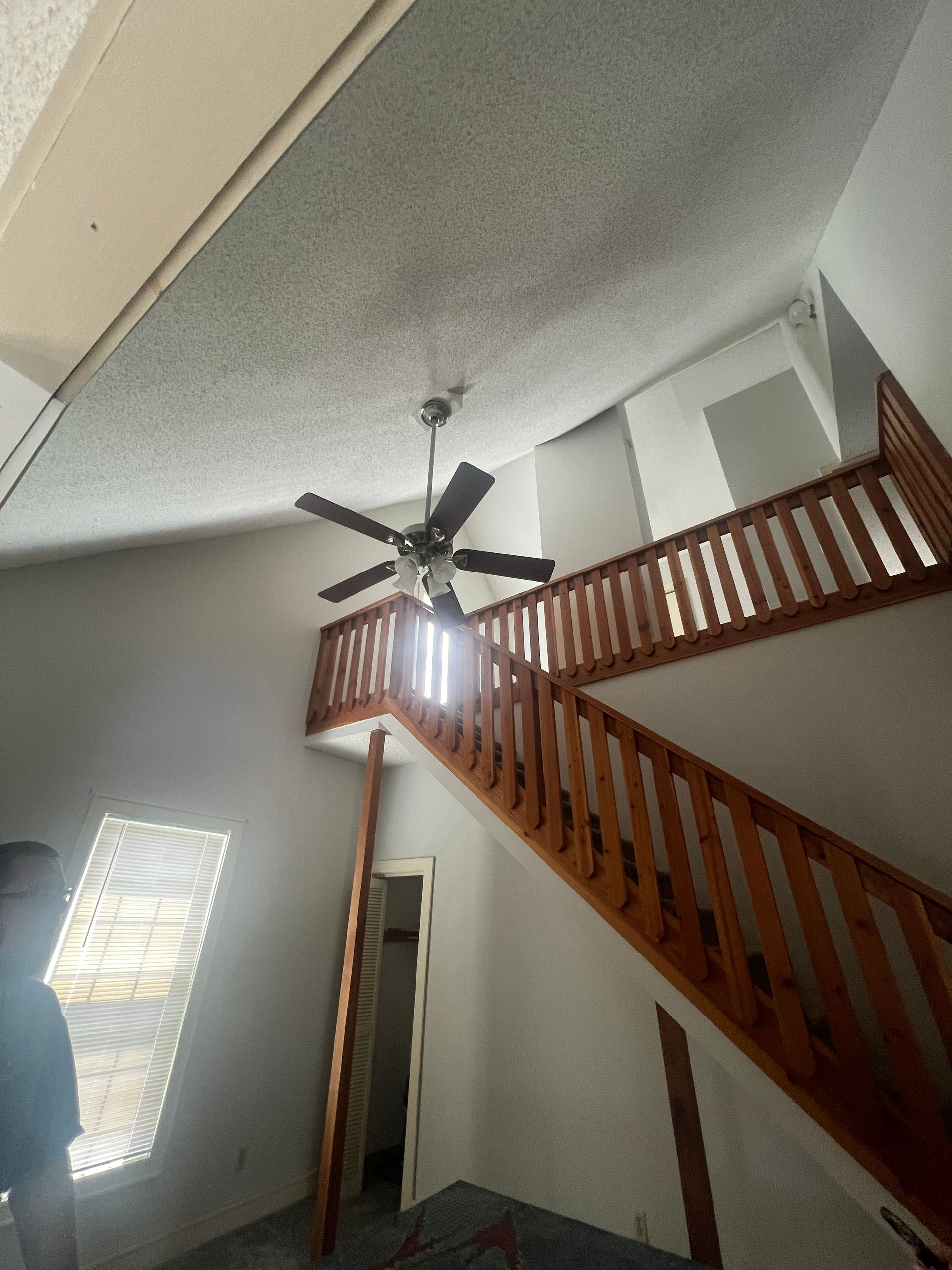 Interior shot of a two-story building with a wooden staircase, high ceiling, and a ceiling fan.