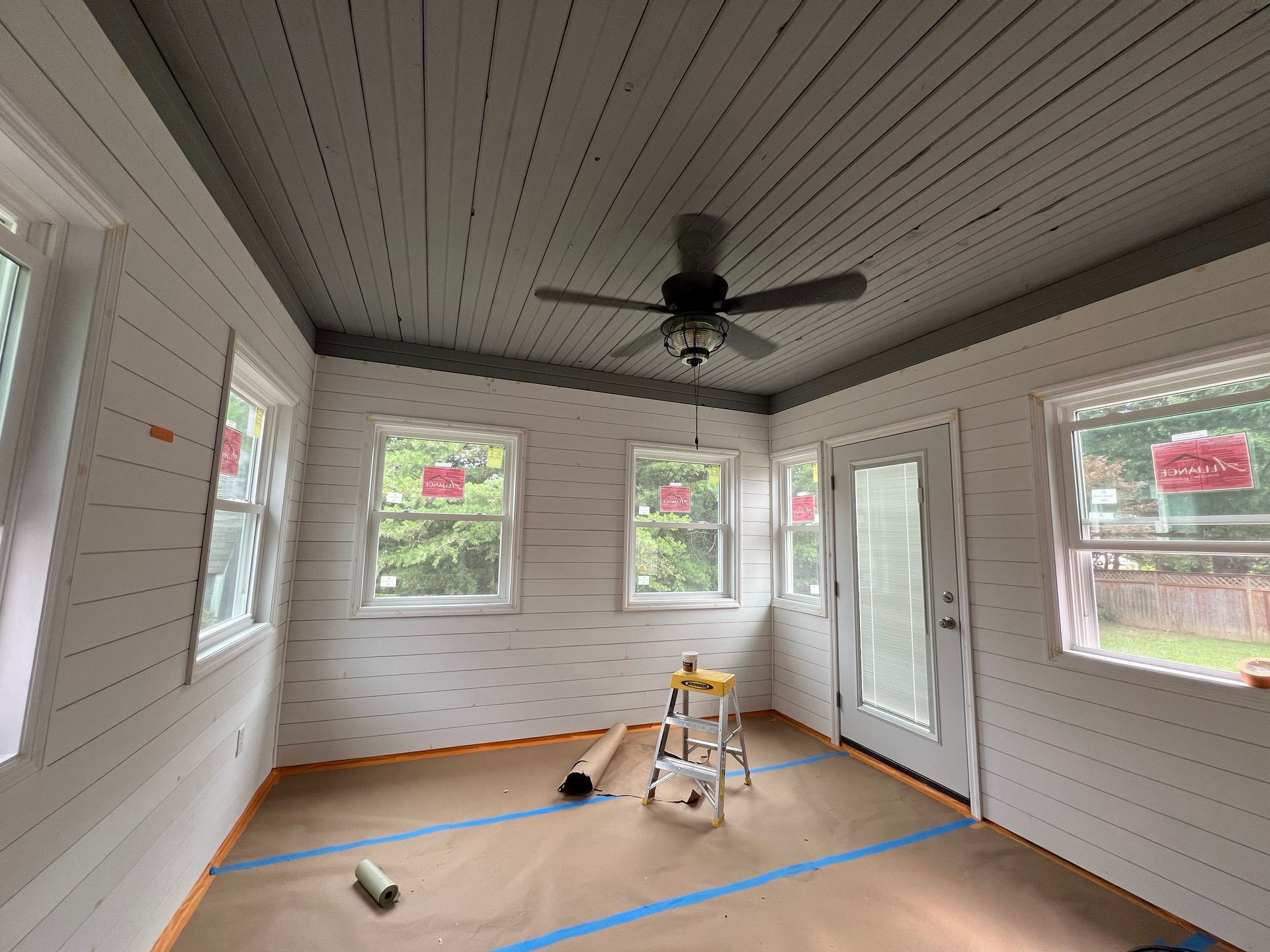 Sunroom interior with white plank walls, gray ceiling, windows, and a ceiling fan.