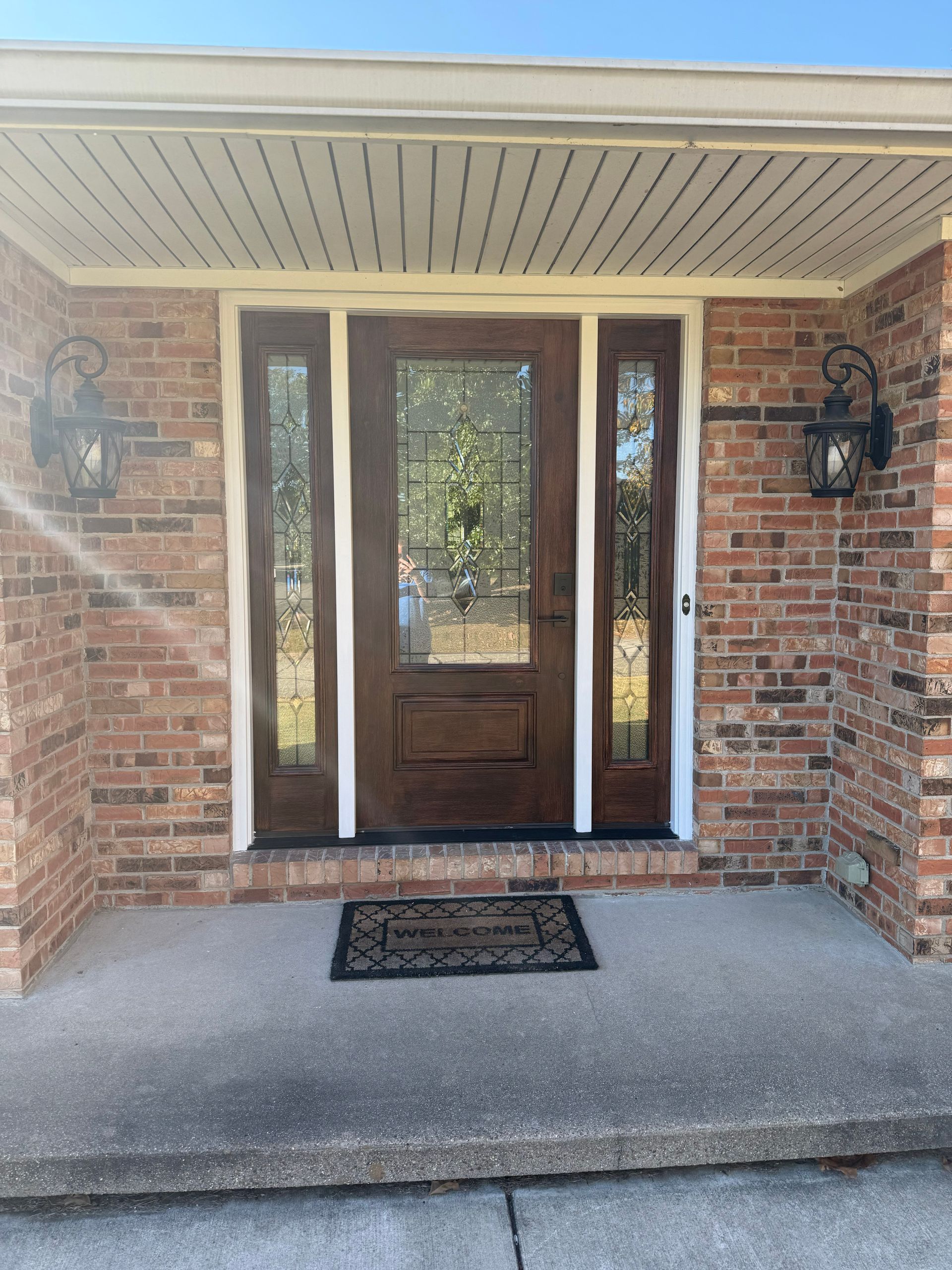 Brick house entrance with brown door, sidelights, and two sconces. Concrete porch with welcome mat.