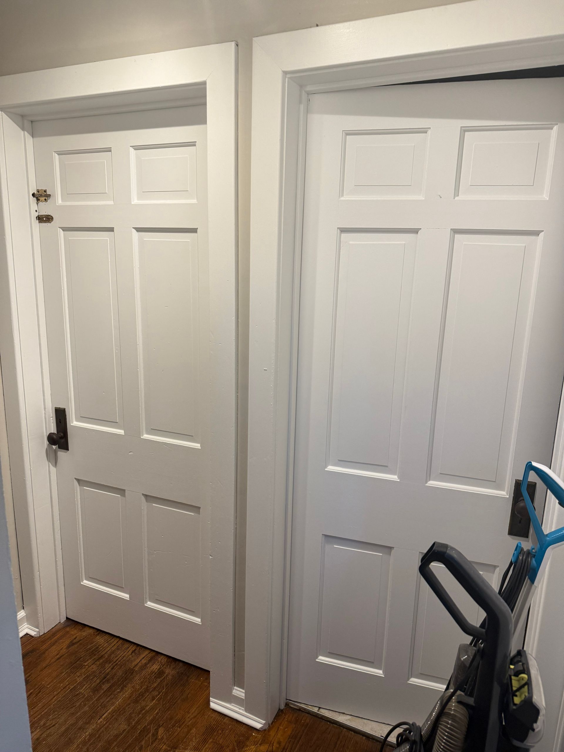 Two white paneled doors side by side. One door is closed; the other is slightly ajar with a vacuum cleaner nearby.