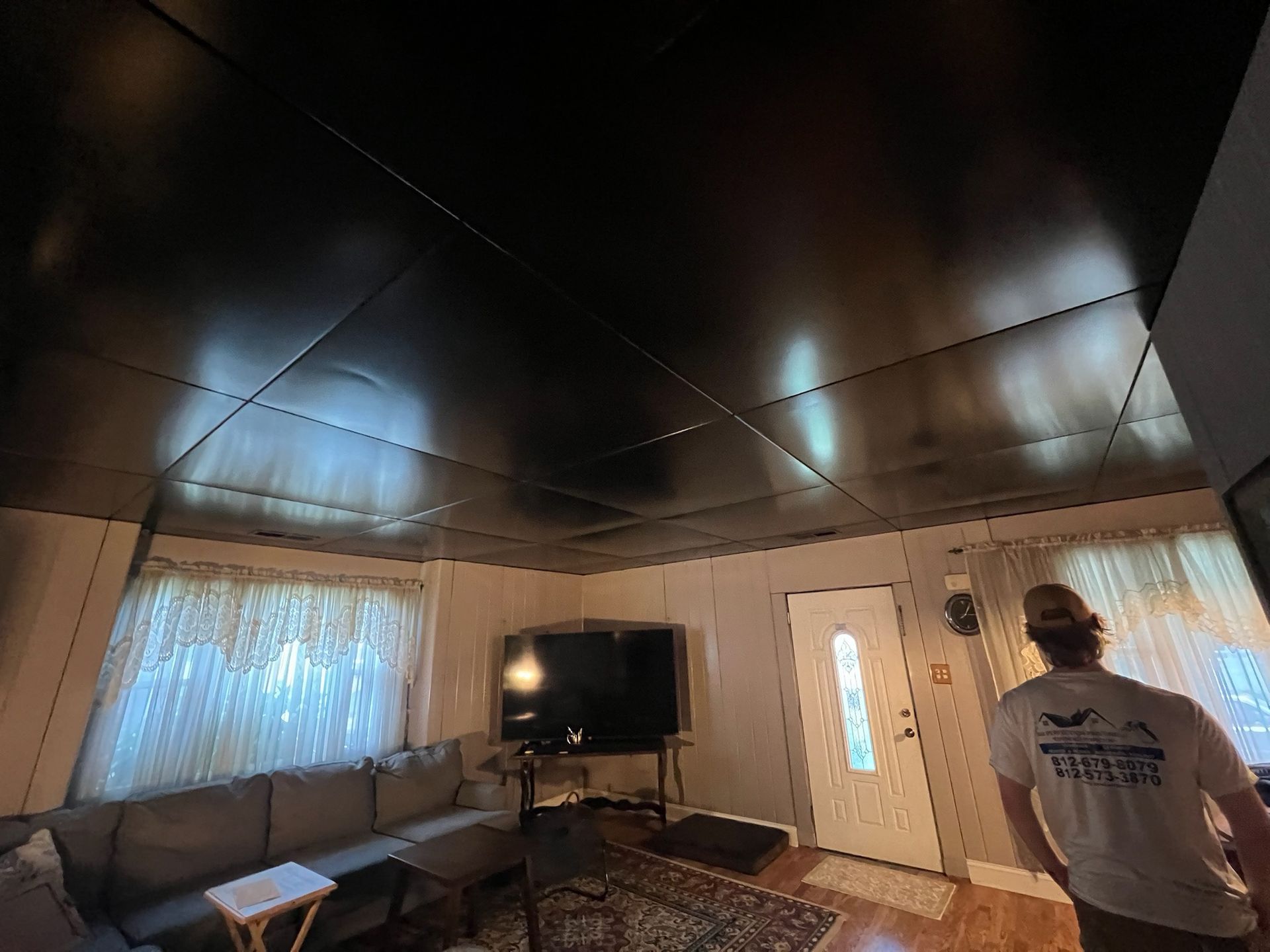 Black ceiling with grid lines, a living room with a couch, TV, door, and a person in a white shirt.