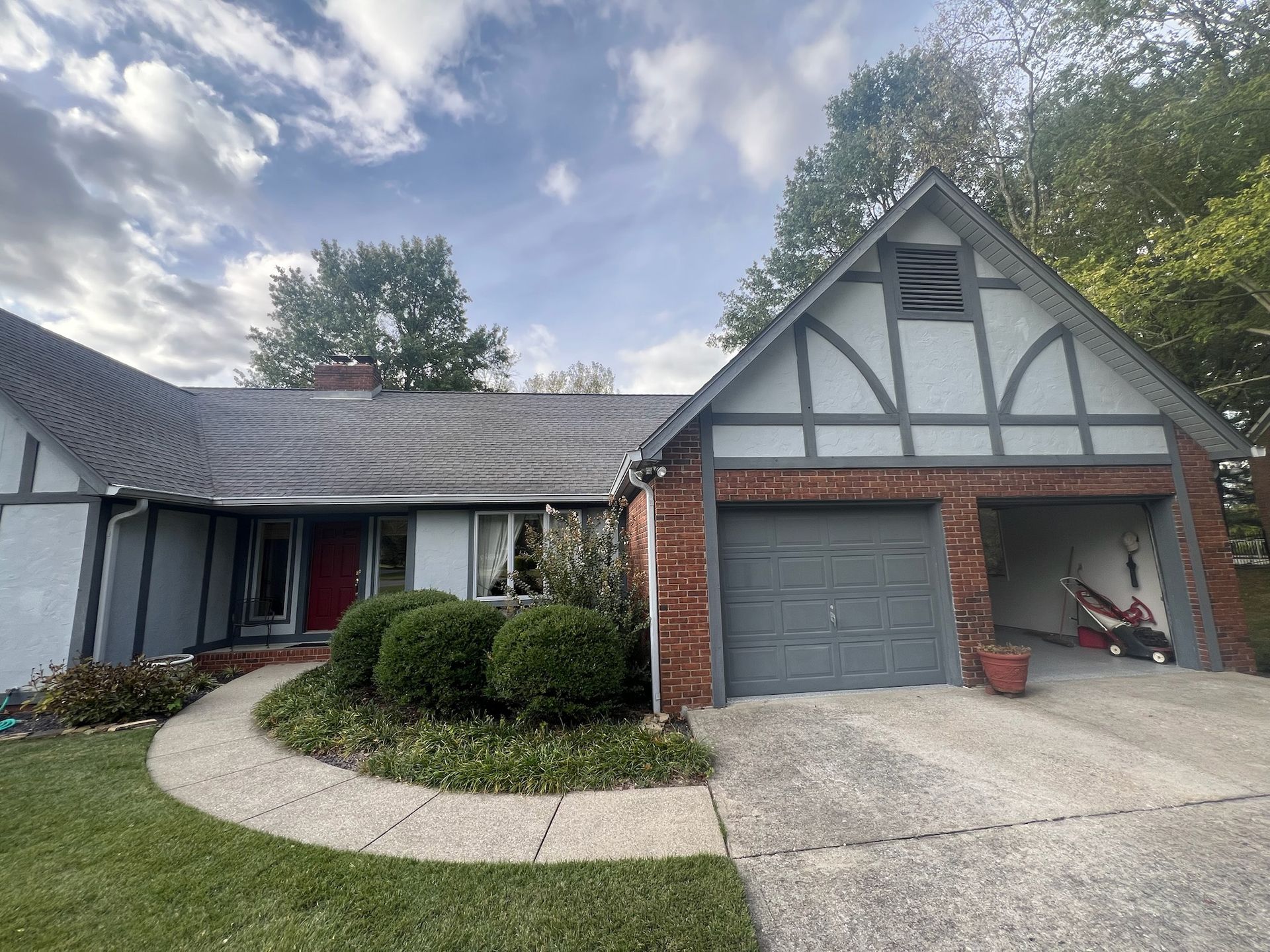 Tudor style house with gray roof, brick garage, and red door. Lush green lawn and sky overhead.
