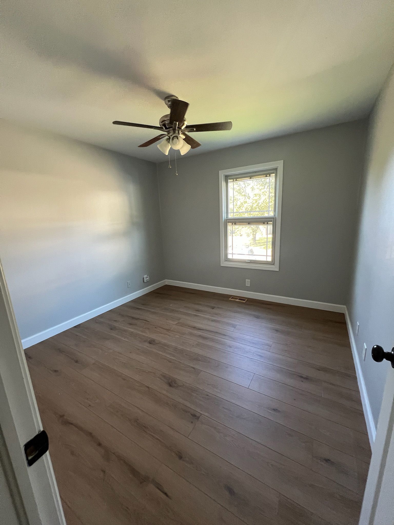 Empty room with gray walls, wooden floor, and a window with sunlight. A ceiling fan hangs overhead.