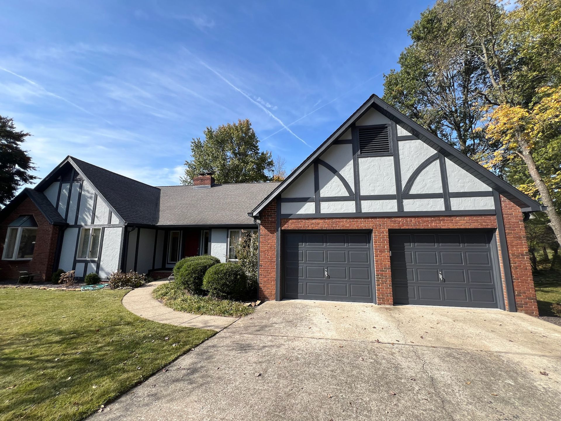 Tudor-style house with red brick, gray trim, and two-car garage. Blue sky, green lawn, and trees.