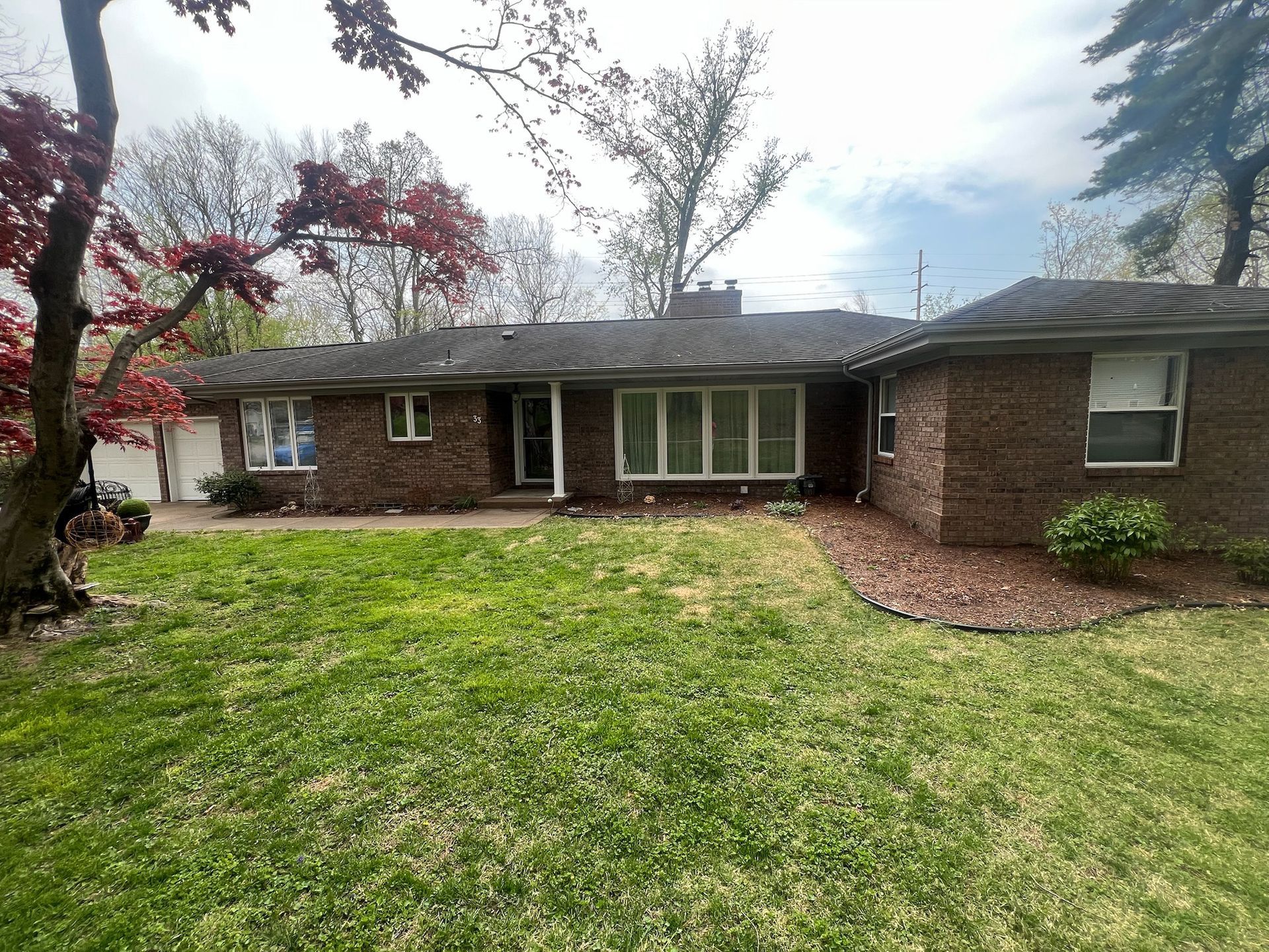 Brick ranch-style house with green lawn and red-leafed tree. Cloudy sky.
