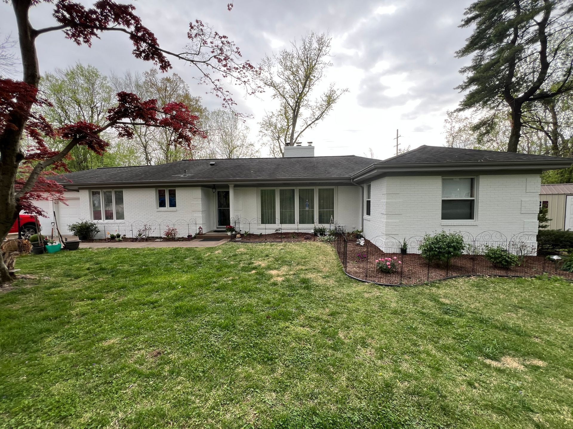 White ranch-style house with a green lawn and trees. Red tree on the left. Cloudy sky.