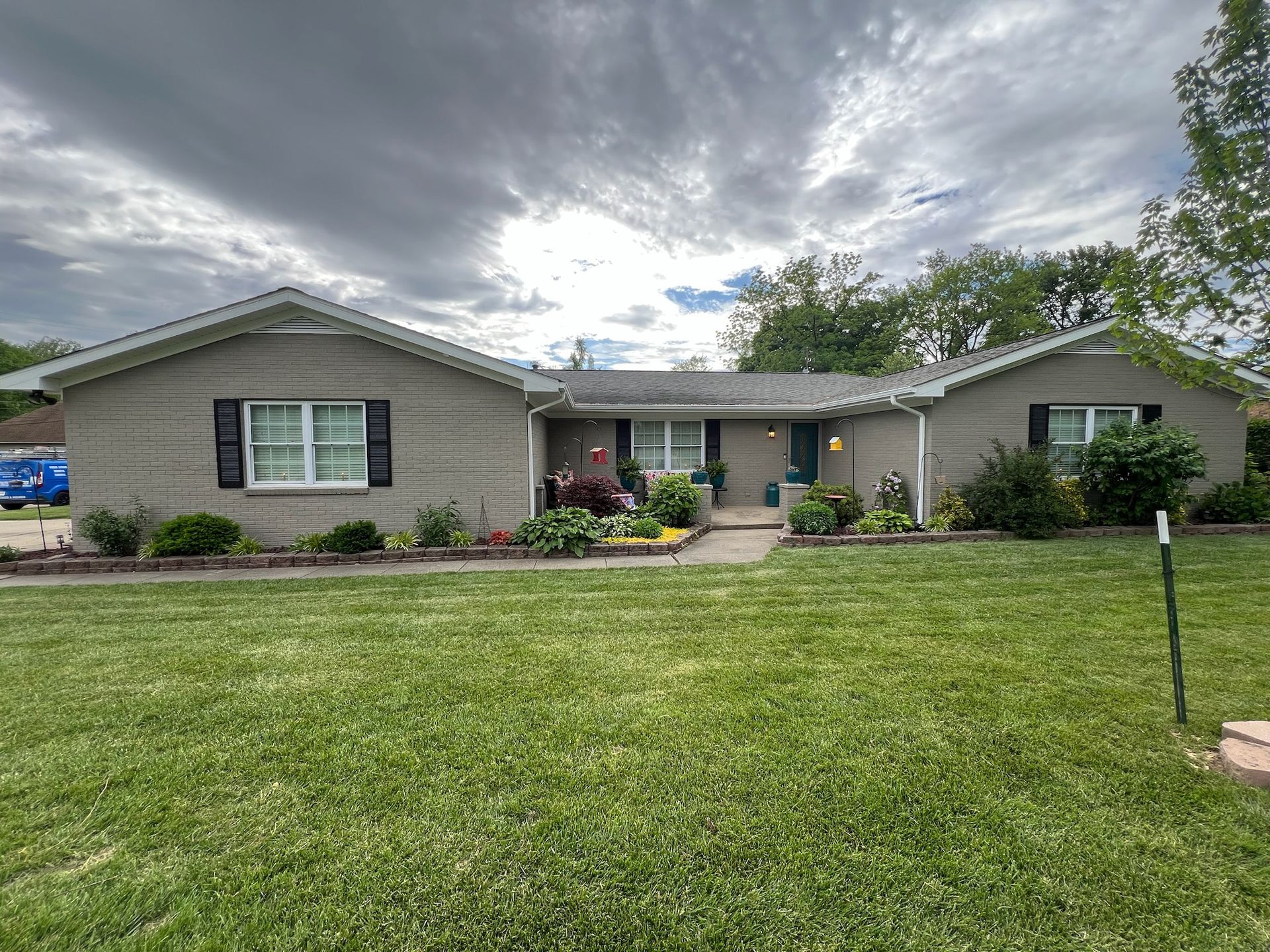 A gray brick ranch house with a green lawn and cloudy sky.