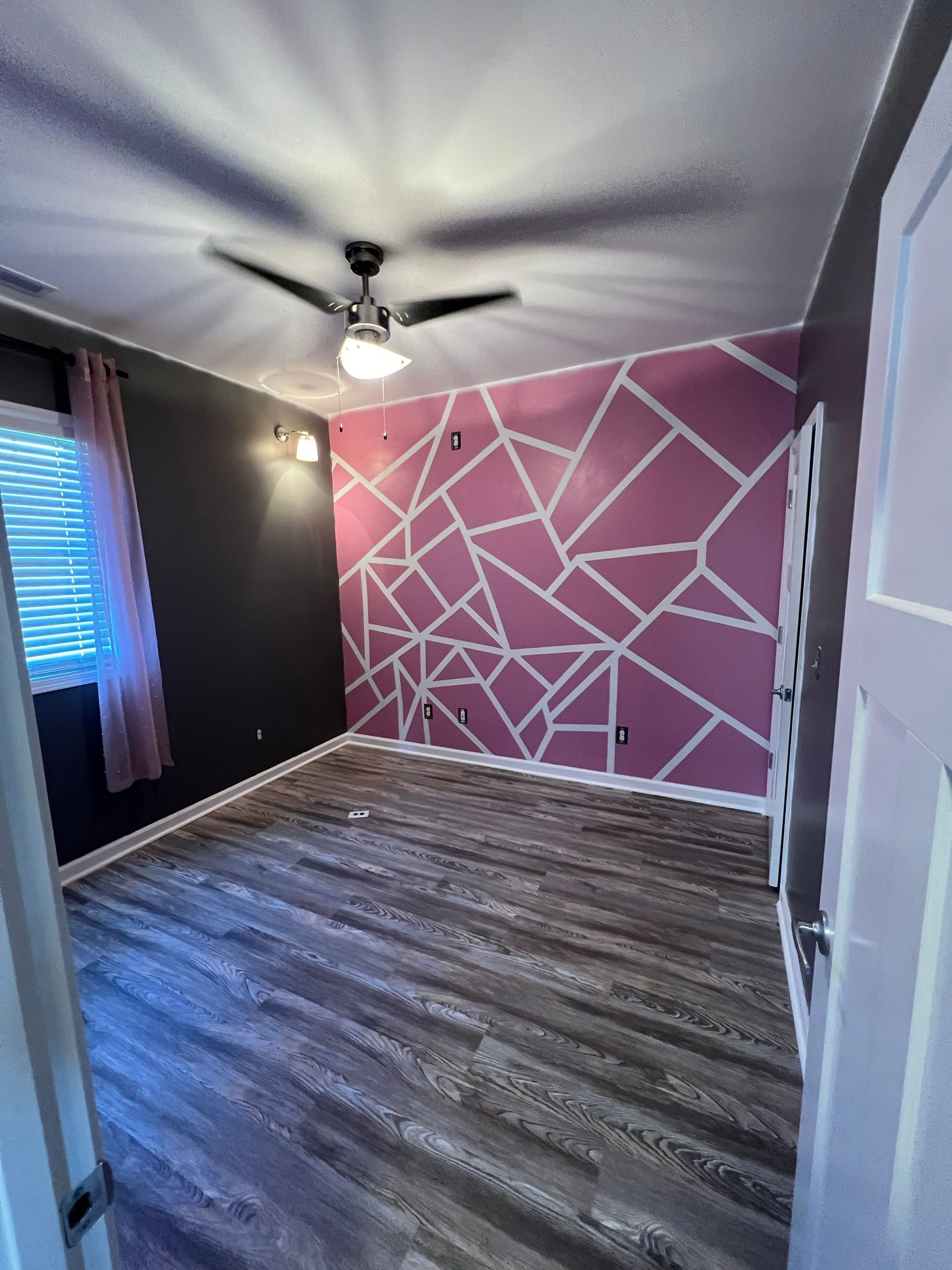 Bedroom with gray wood floors, pink accent wall with white geometric design, dark gray wall, and a ceiling fan.
