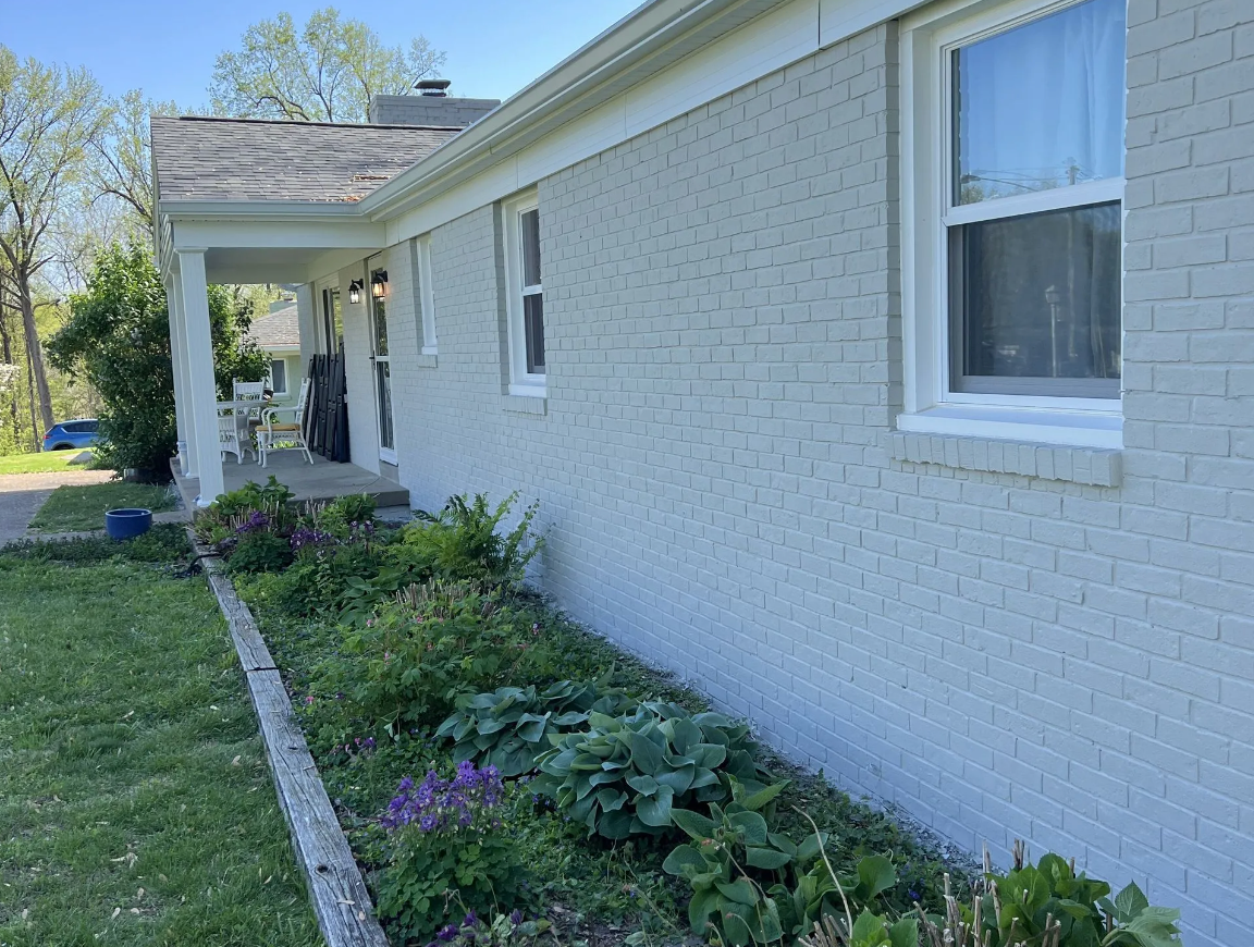 Gray-painted brick house with white trim, windows, and porch. A flowerbed with plants sits in front of the house.