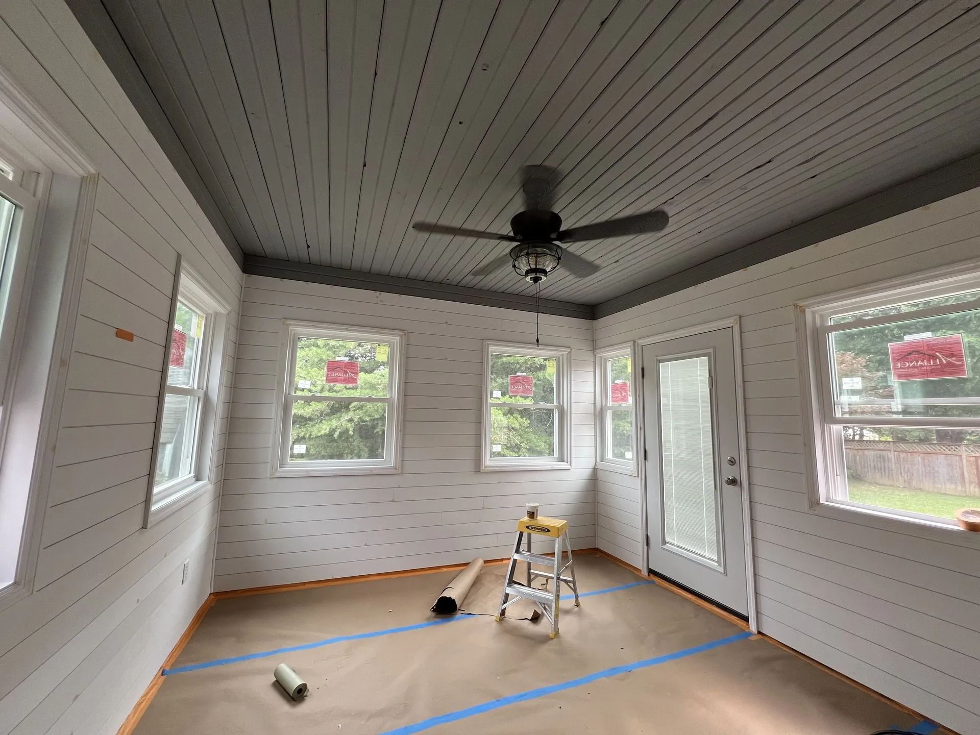 Sunroom interior with white shiplap walls, gray ceiling, windows, a ceiling fan, and a door.