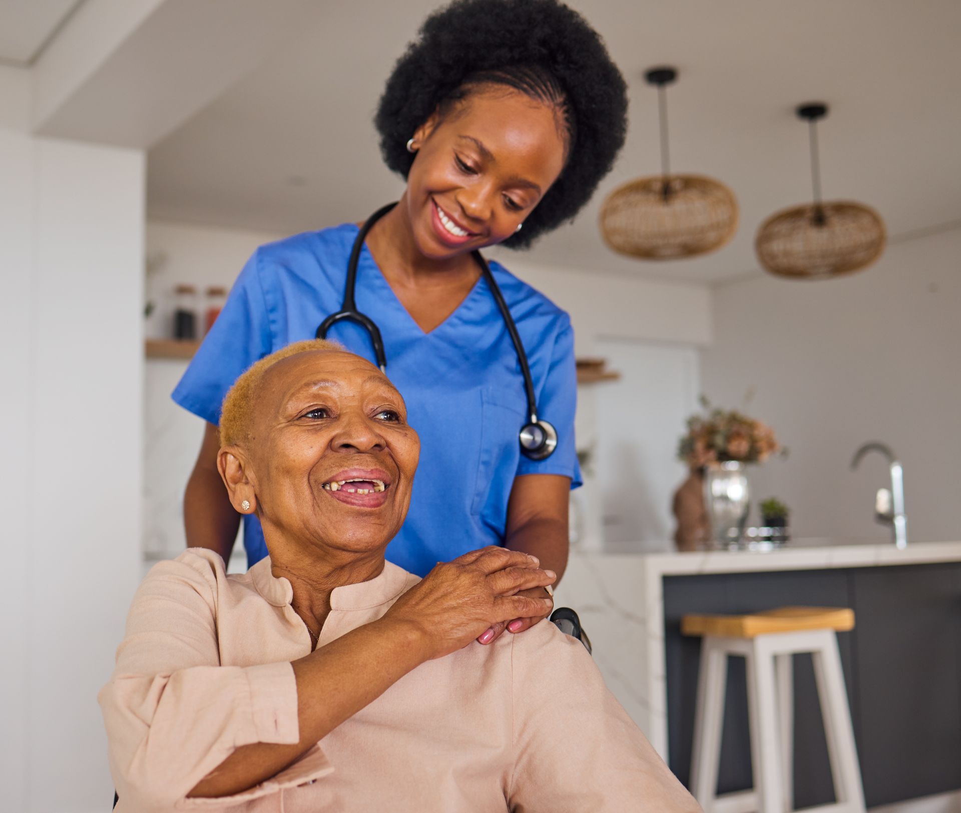 Smiling person in blue scrubs with stethoscope, assisting a person in a wheelchair indoors.