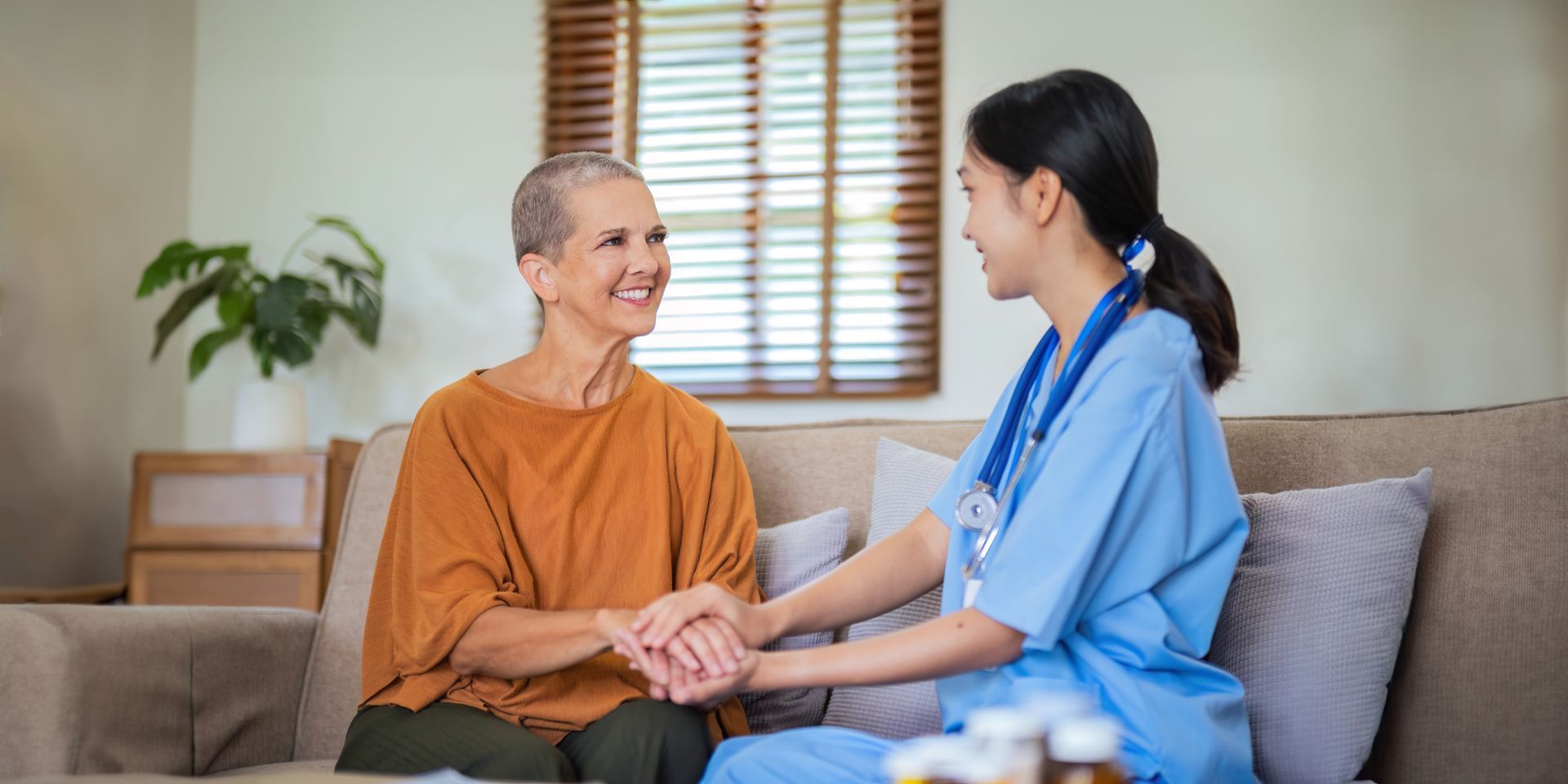 A caregiver in blue scrubs holding hands with a woman in an orange shirt. They are seated on a couch.