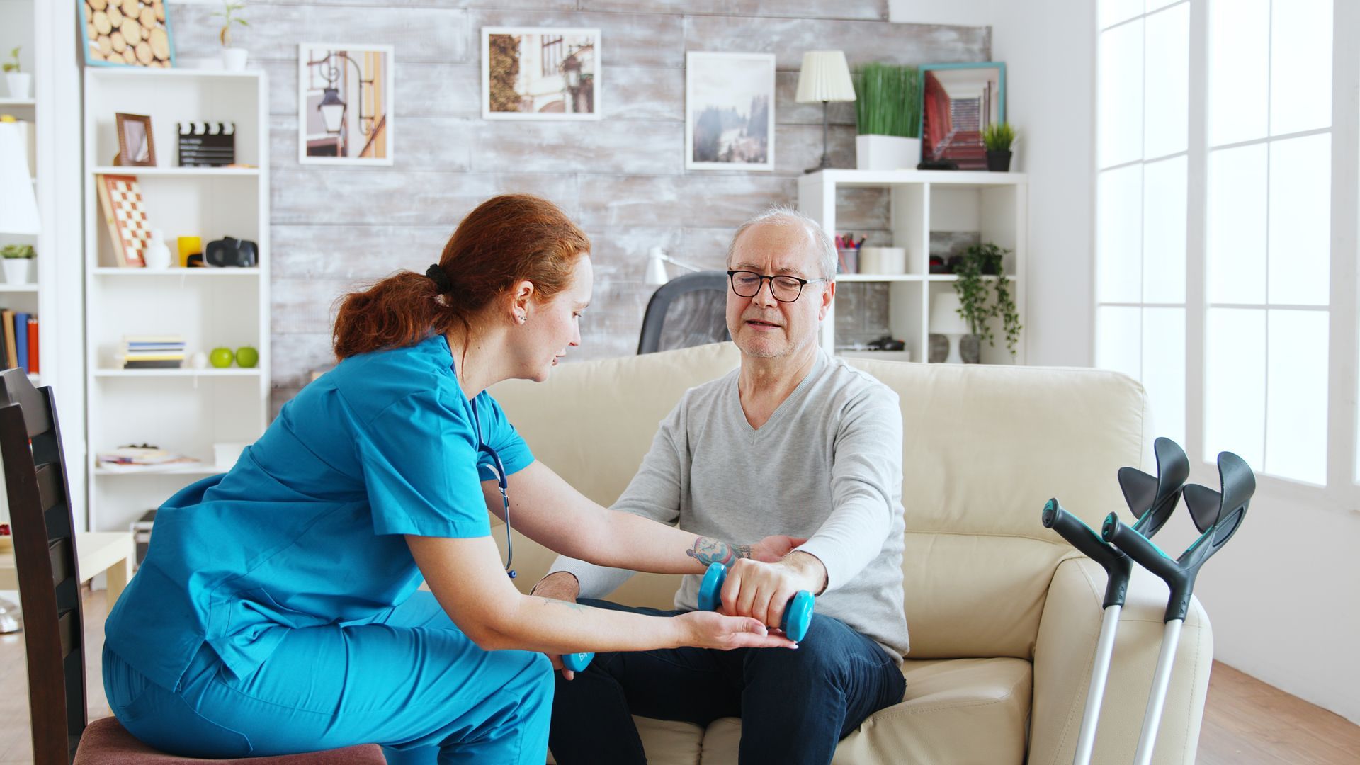 Caregiver assisting a person with arm exercise using a dumbbell. Indoor setting, sofa.