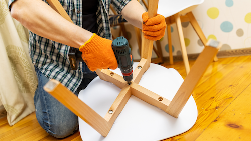 Person in orange gloves uses a power drill to assemble a wooden chair, indoors.