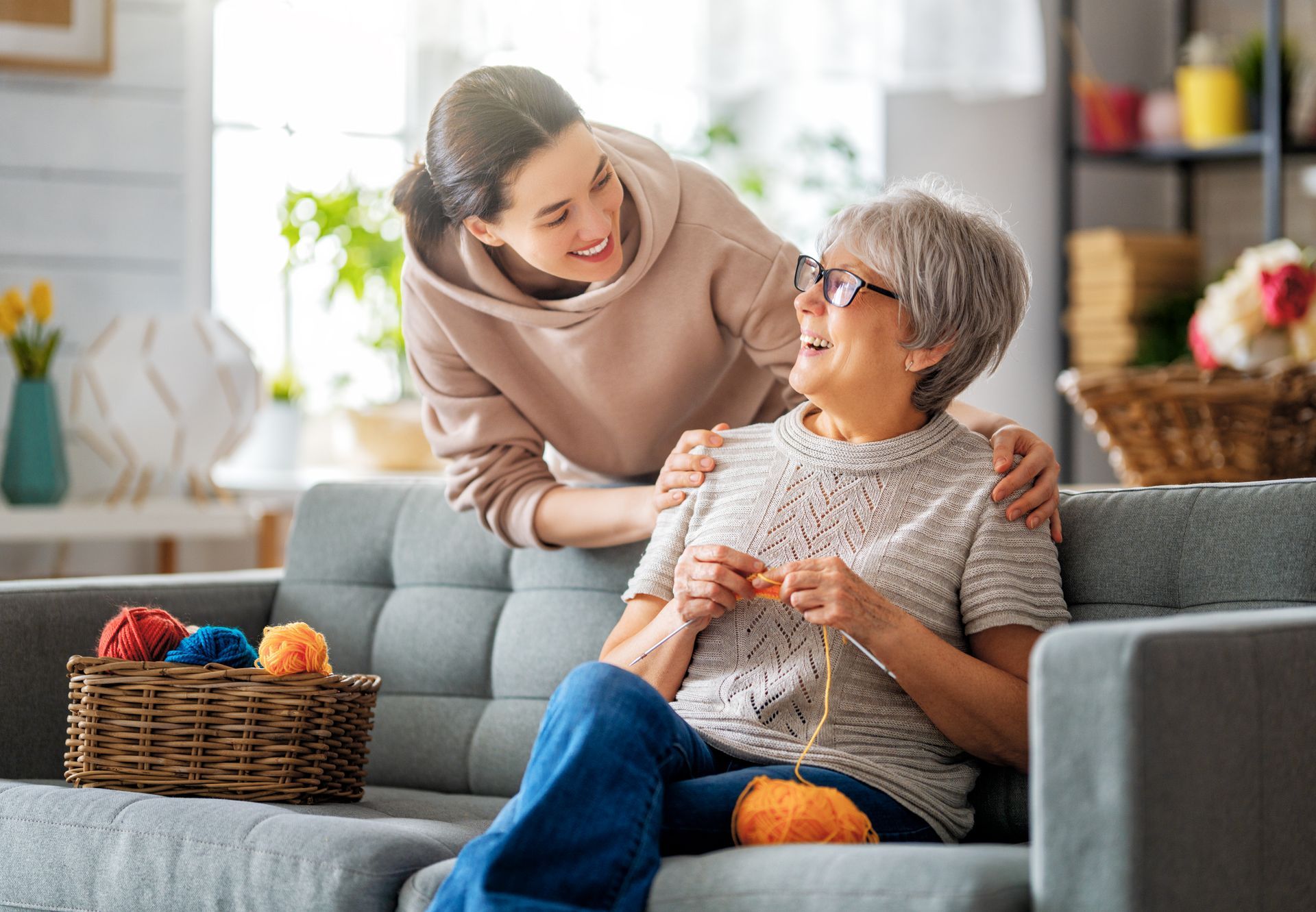Woman with arm around woman knitting, both smiling on a sofa. Basket of yarn nearby.
