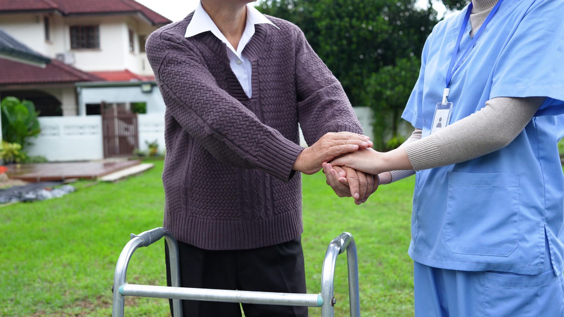Person using a walker assisted by someone wearing scrubs in a grassy yard.