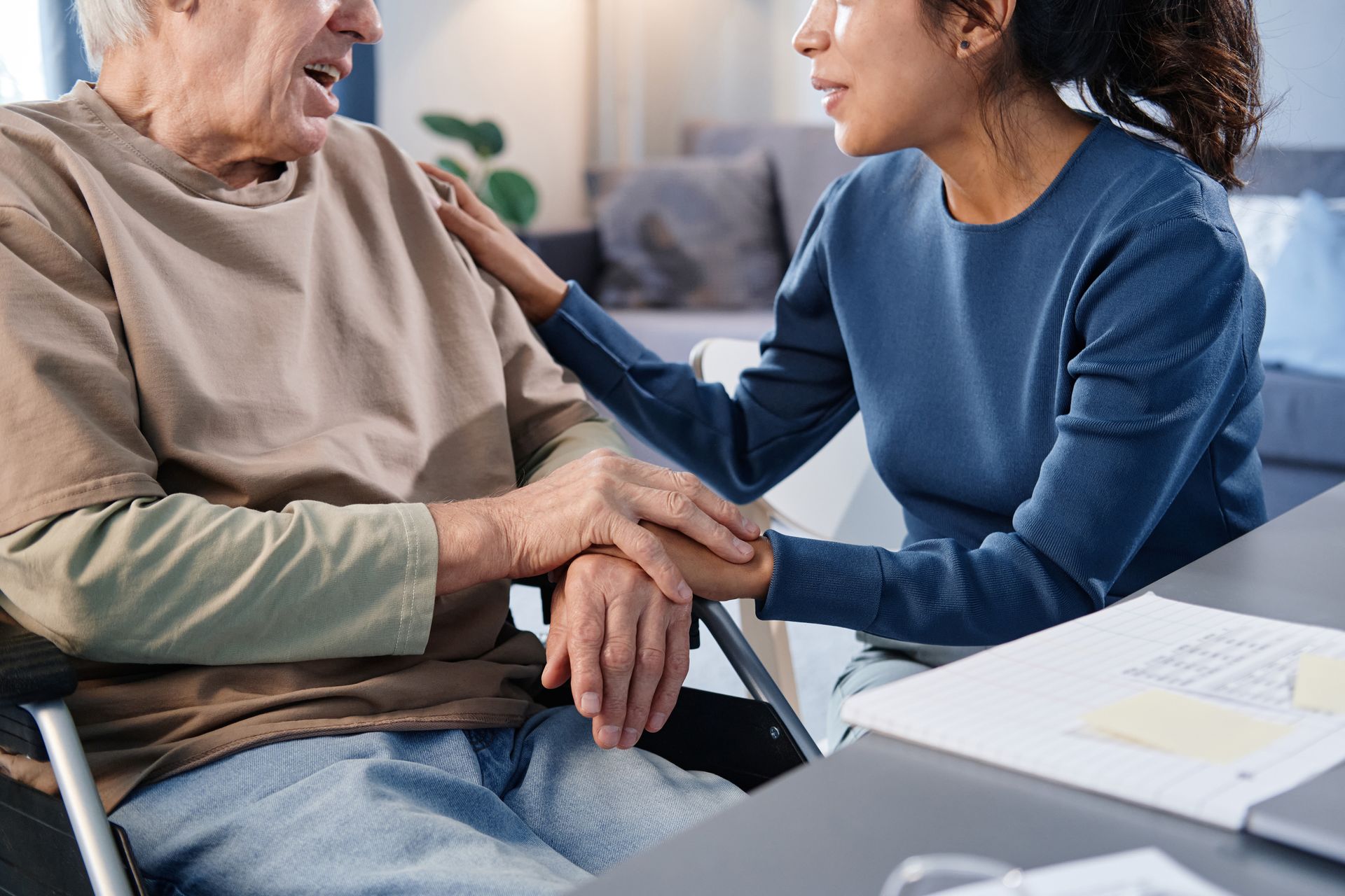 Woman comforts man in wheelchair, hand on his arm and shoulder, interior setting.
