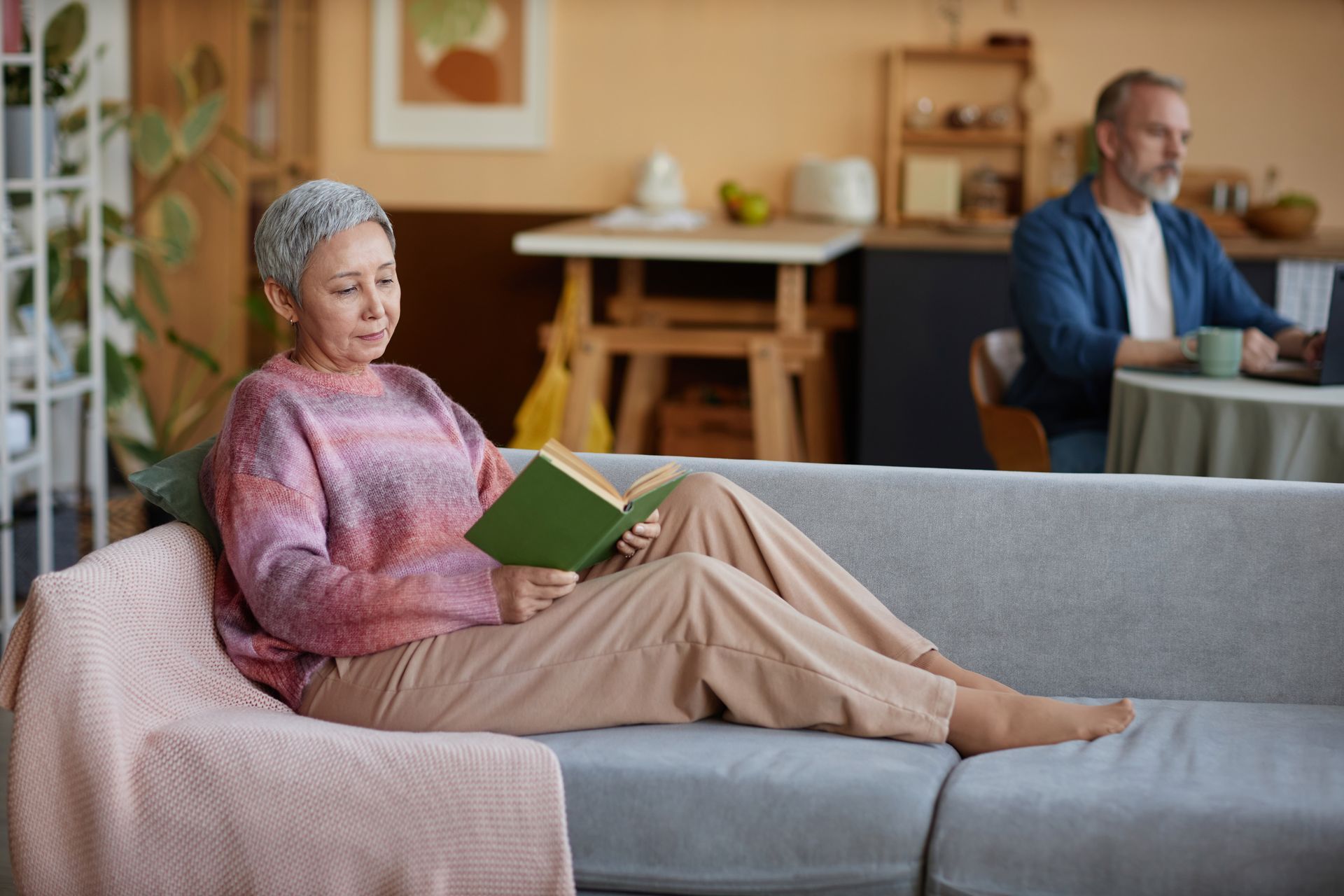 Woman reading book on a couch; man works on laptop in background. Living room with plants, neutral colors.