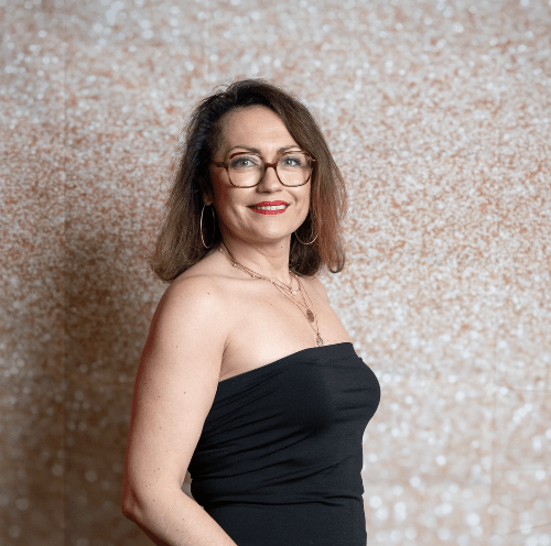 Une femme portant des lunettes et une robe noire se tient devant un mur.