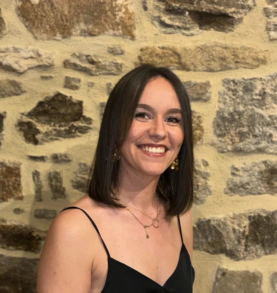 Une femme en robe noire sourit devant un mur de pierre.