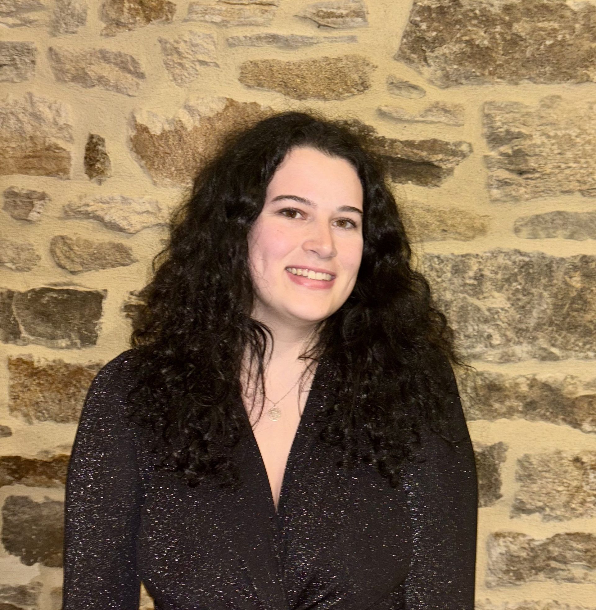 Une femme en robe noire se tient devant un mur de pierre.