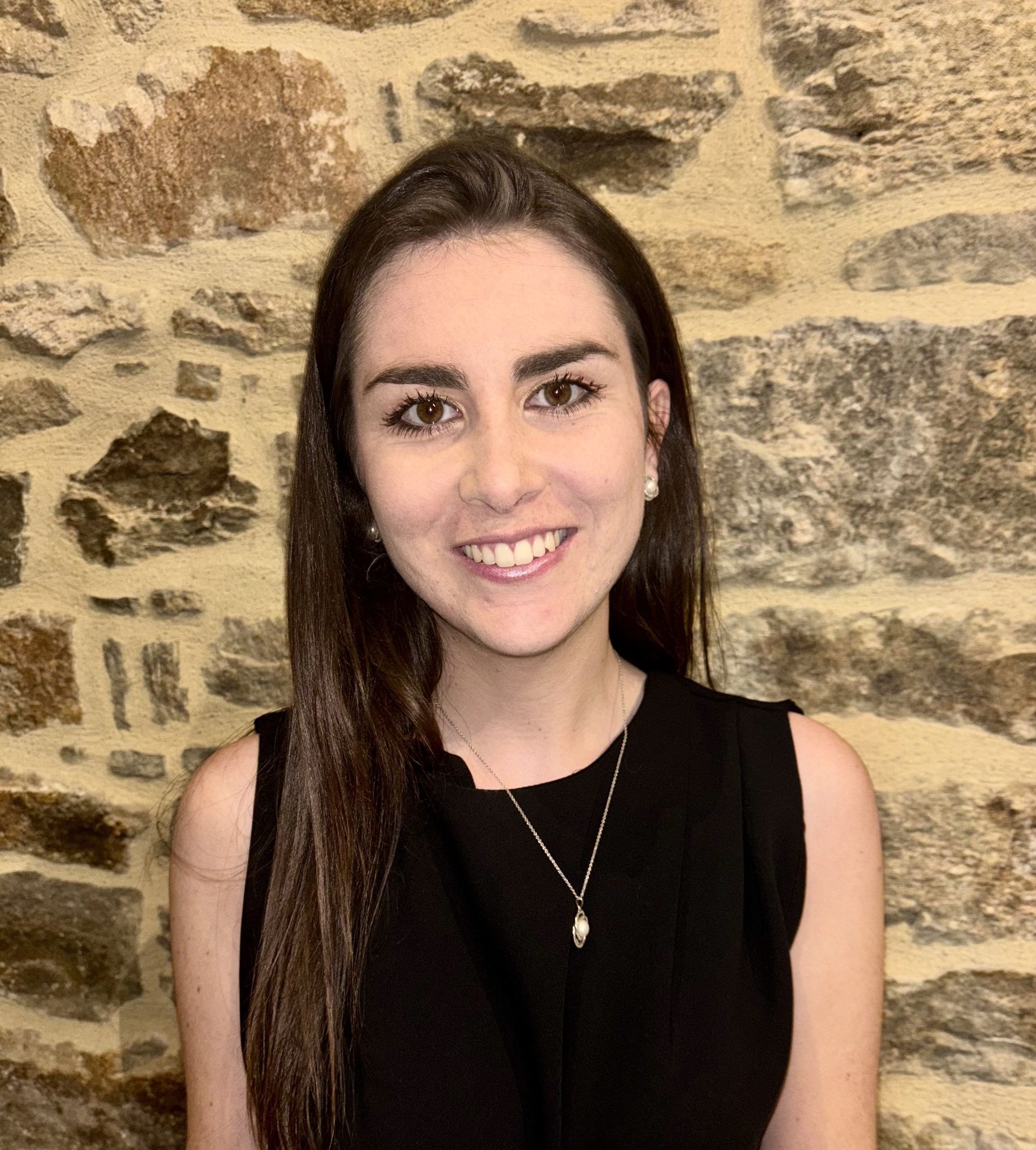 Une femme en robe noire sourit devant un mur de pierre.