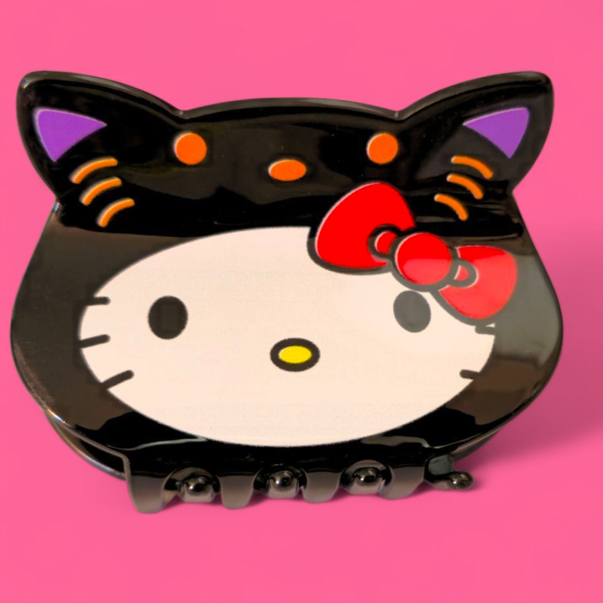 Pinza para el pelo Hello Kitty negra con orejas moradas, detalles naranjas y lazo rojo.