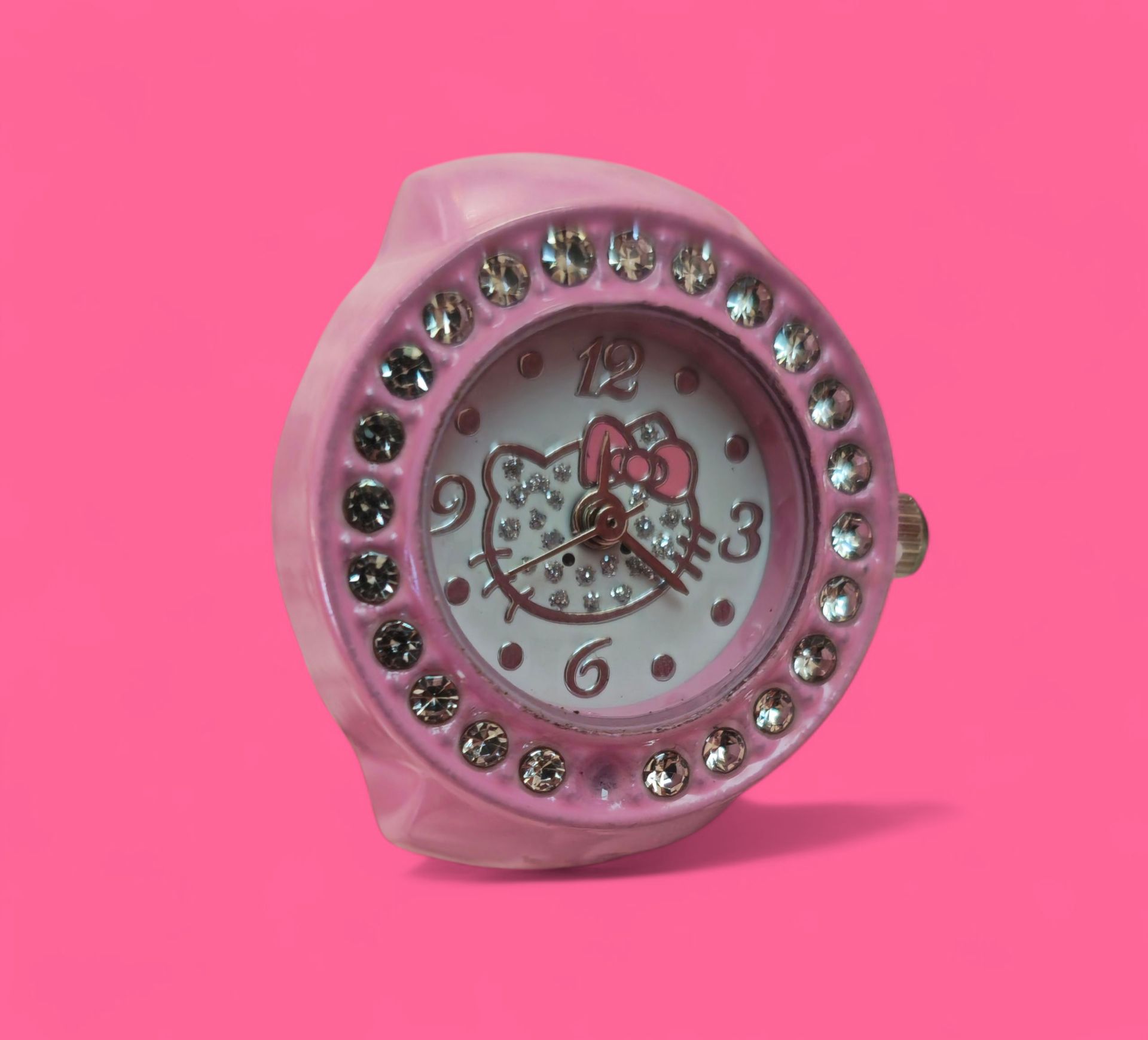 Reloj de pulsera tipo anillo Hello Kitty rosa con bisel con incrustaciones de joyas sobre fondo rosa.