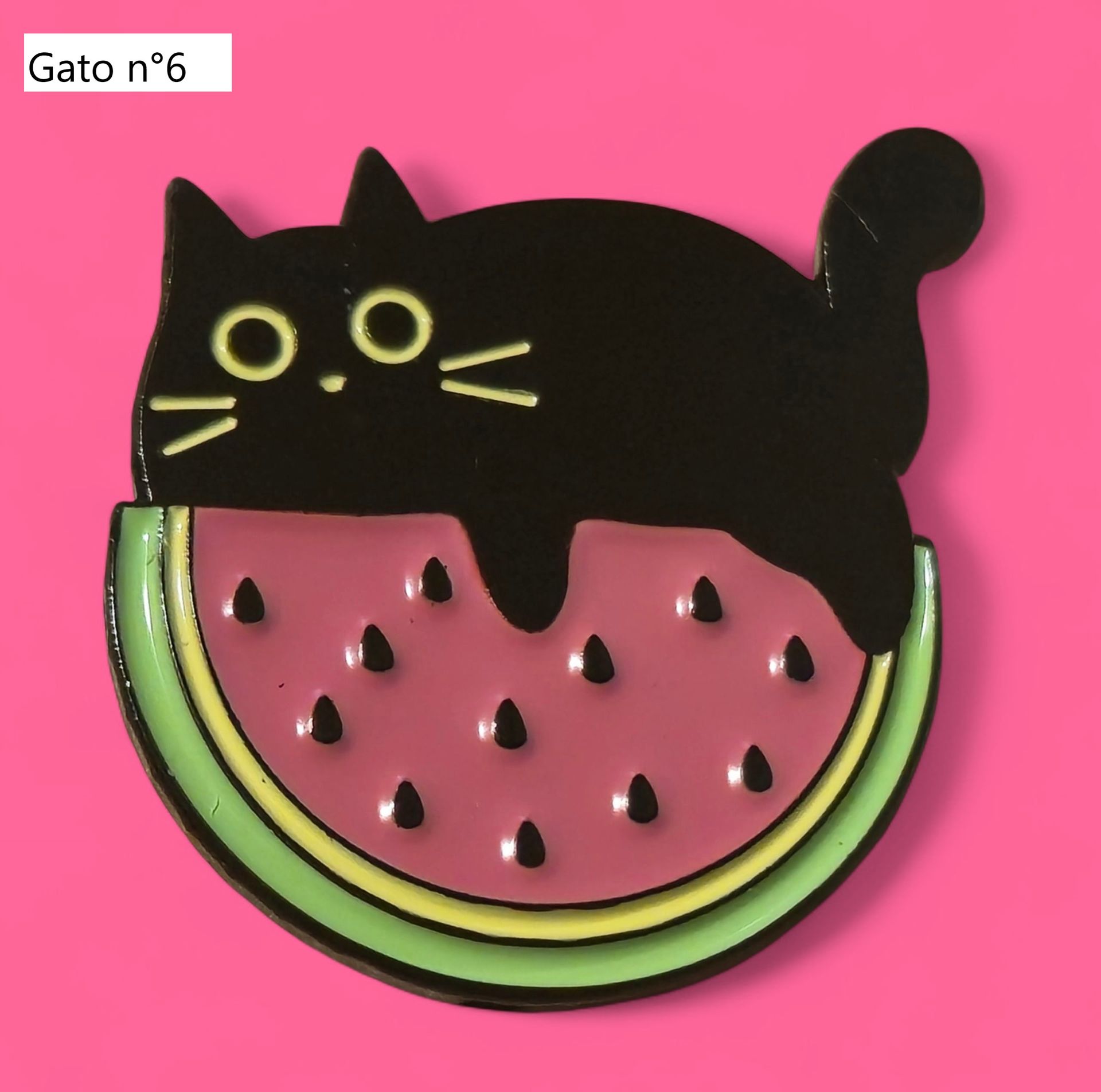 Pin de esmalte de gato negro con ojos dorados, rodeado de hojas de plantas verdes, sobre un fondo rosa.