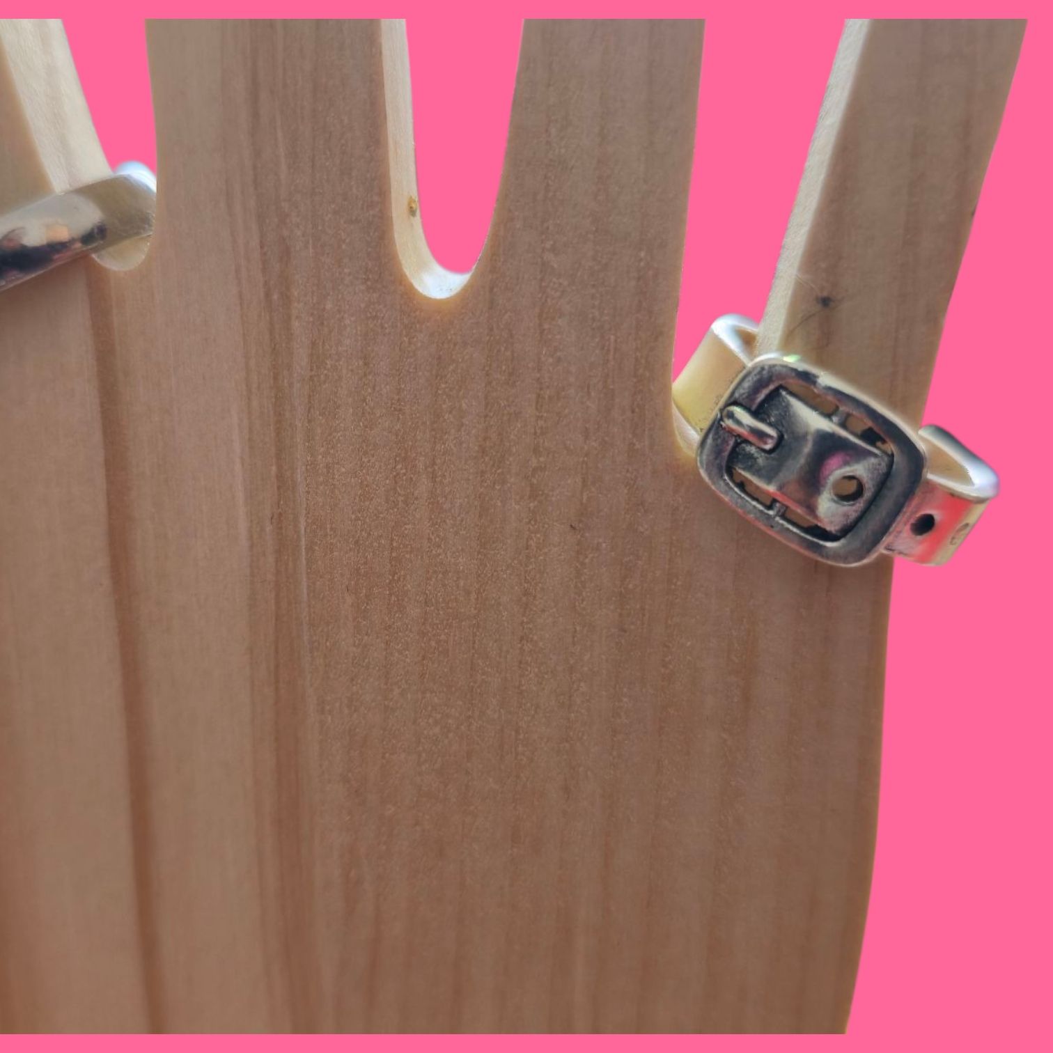 Anillo plateado con diseño de hebilla sobre una mano de madera con fondo rosa.