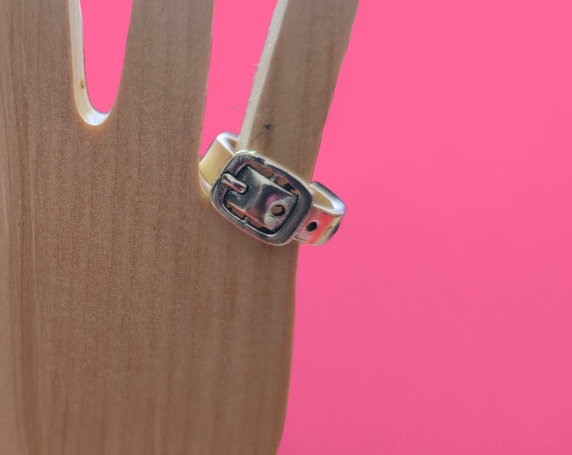 Anillo plateado con diseño de hebilla sobre una mano de madera con fondo rosa.