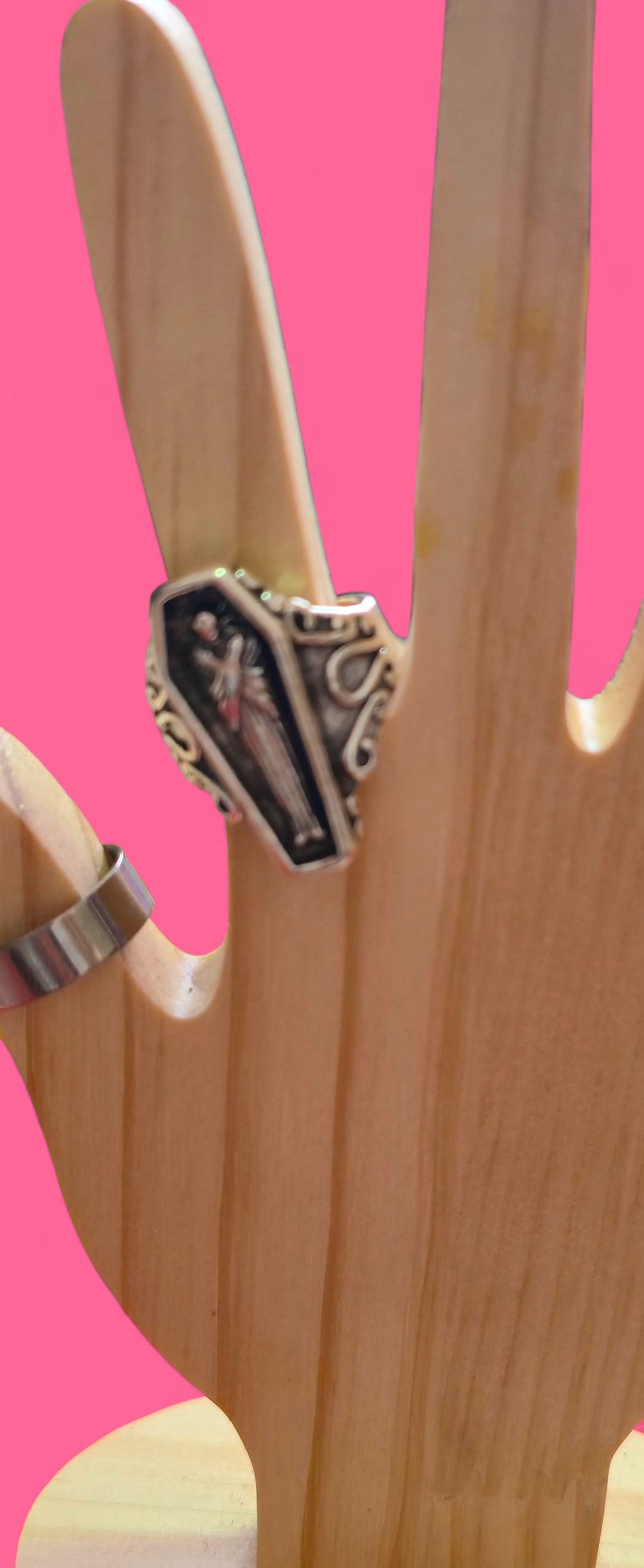 Anillo con diseño de ataúd sobre un soporte de madera con fondo rosa.