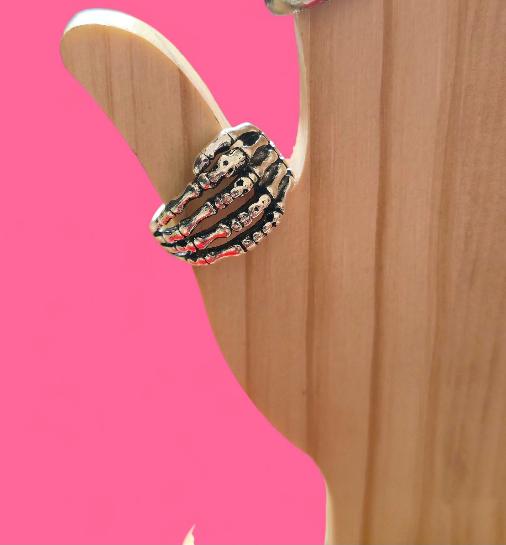 Anillo de plata con forma de mano de esqueleto sobre un soporte de madera, fondo rosa.
