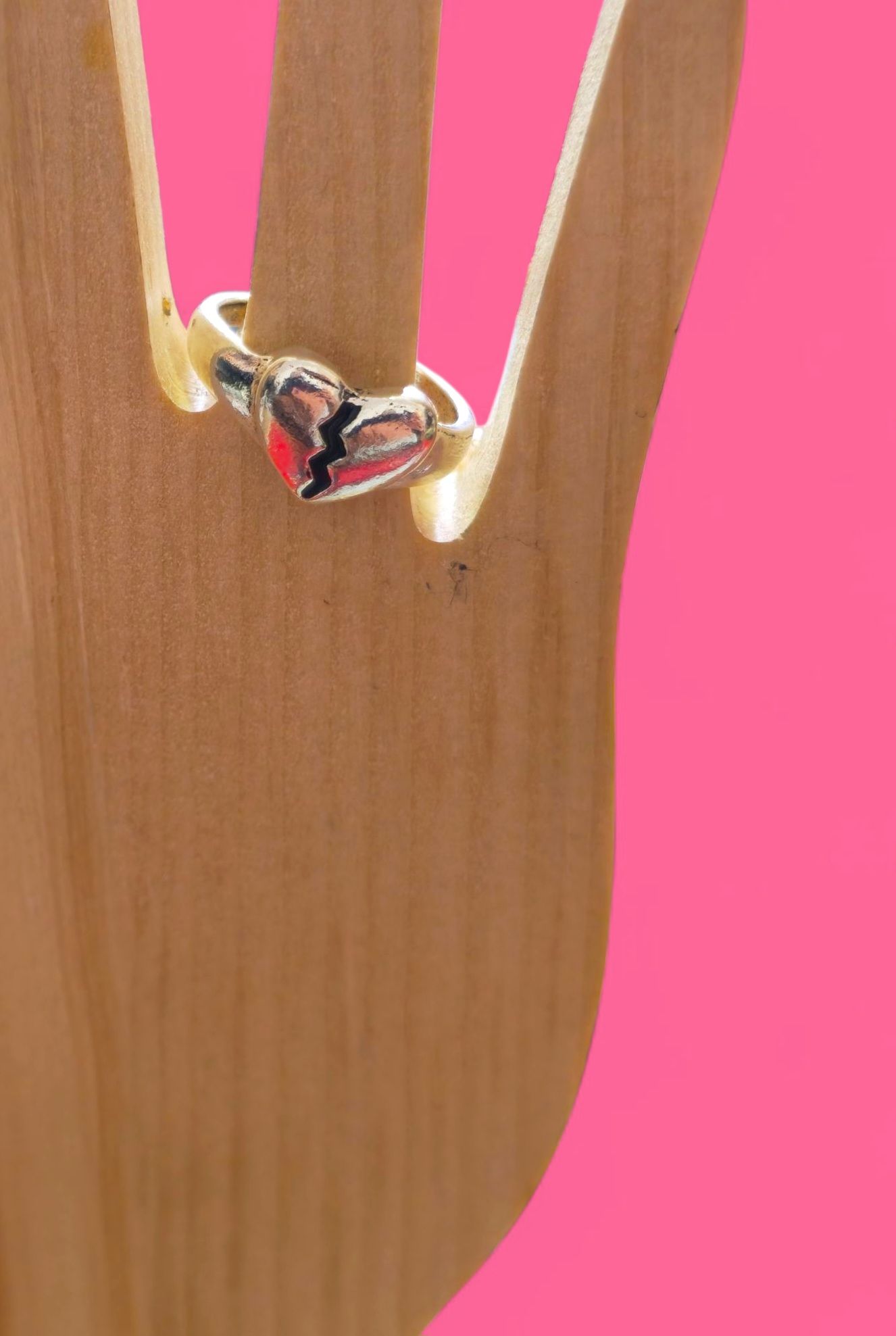 Anillo de plata con un corazón rojo agrietado sobre una mano de madera, sobre un fondo rosa.