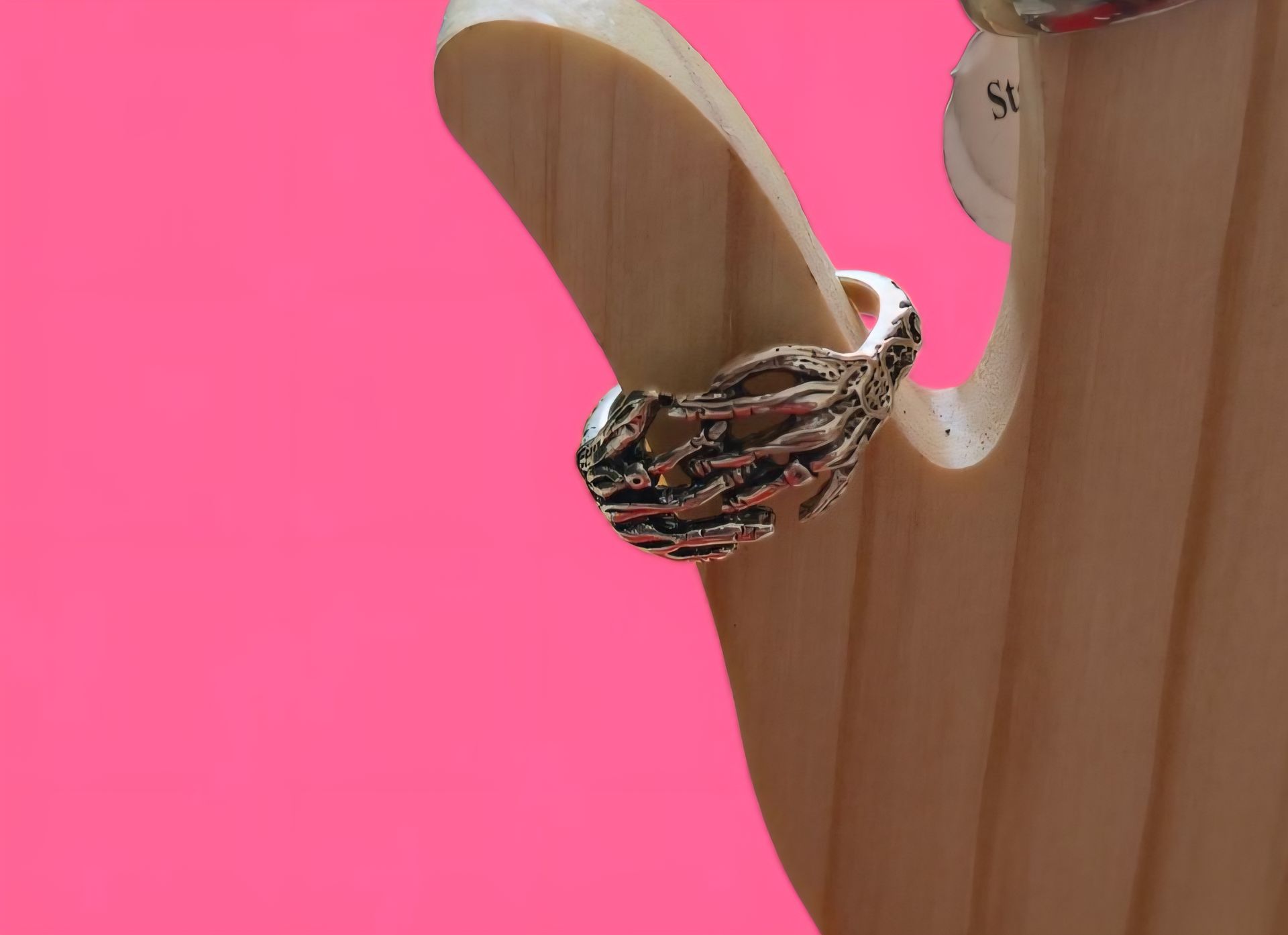 Anillo de plata con forma de mano de esqueleto sobre un soporte de madera, fondo rosa.